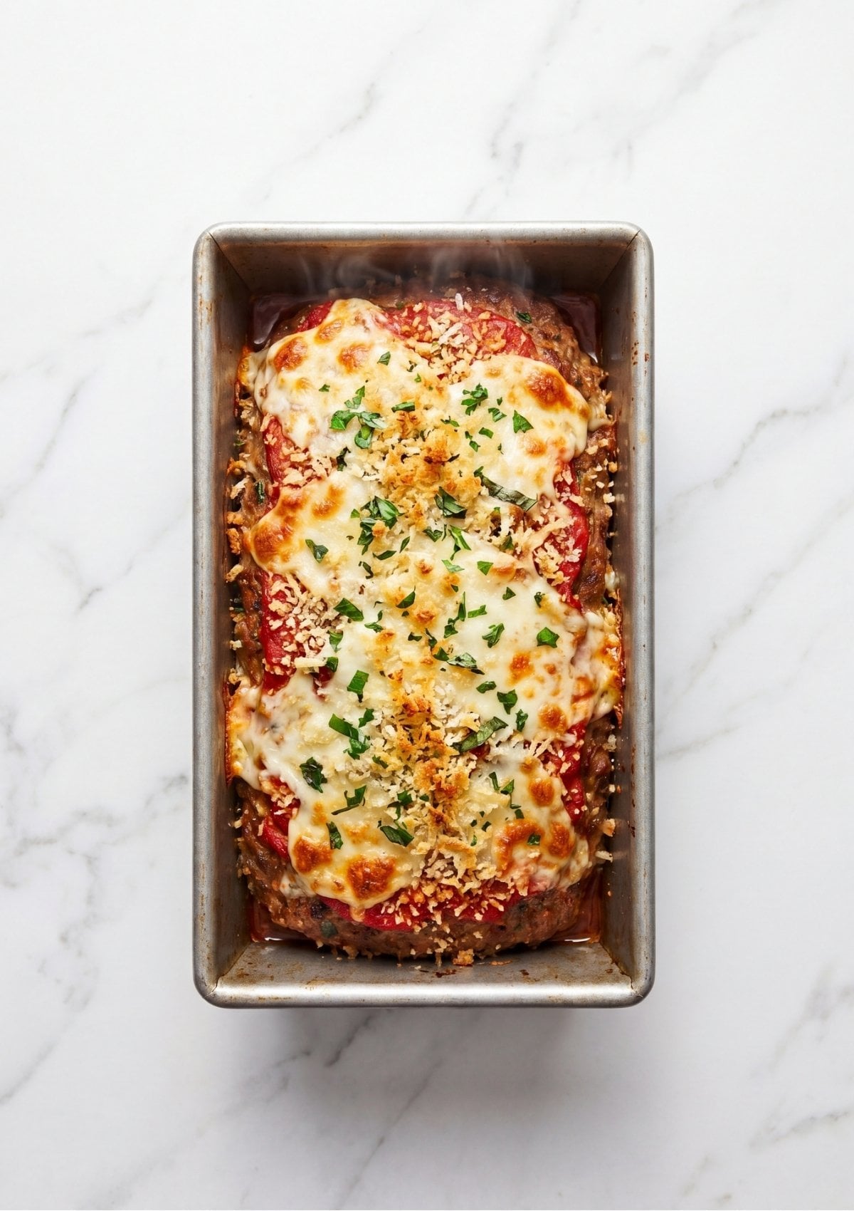 A freshly baked Italian meatloaf still in the loaf pan, featuring a golden-brown crust of melted cheese, herbs, and crispy breadcrumbs.