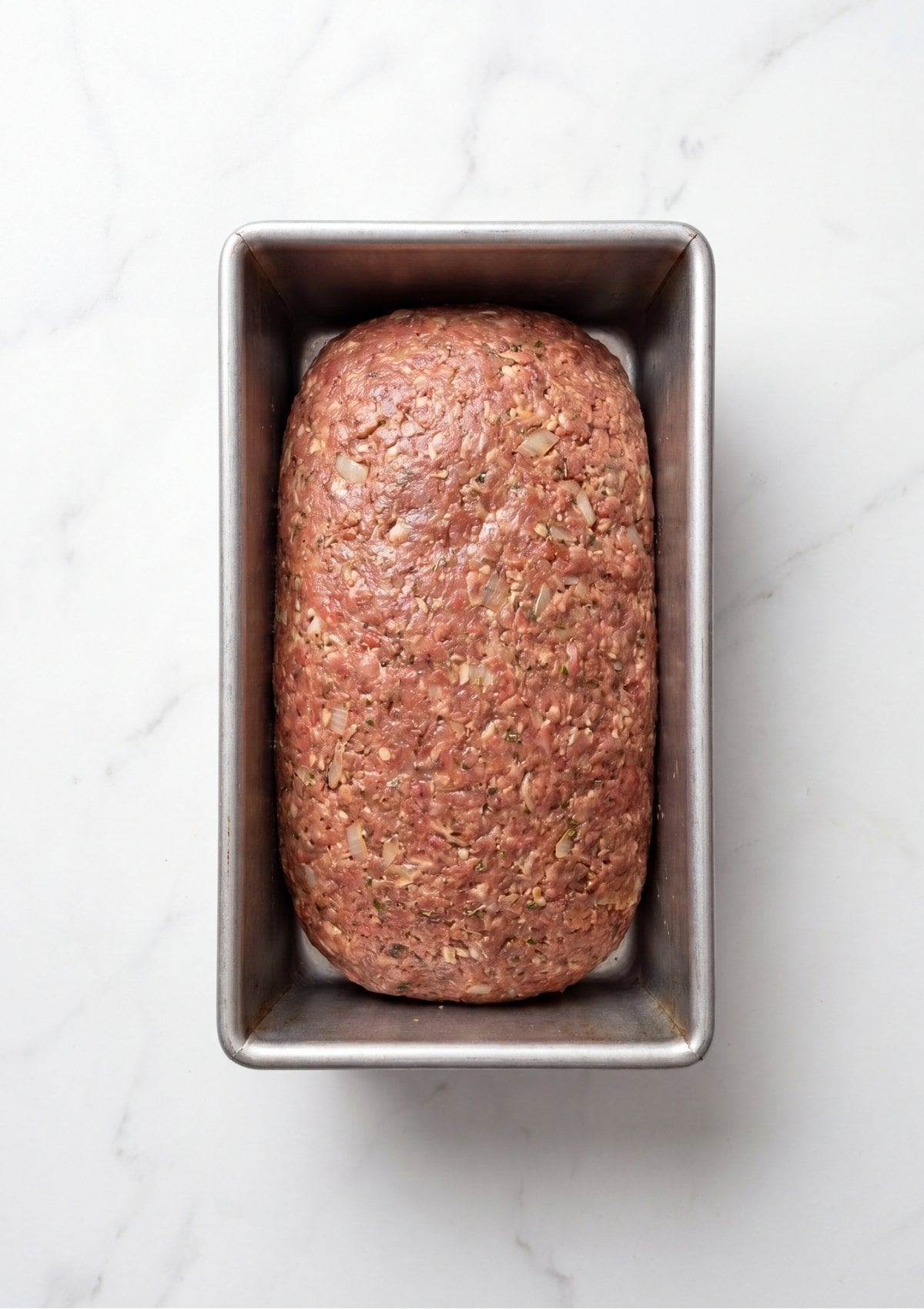 A raw, seasoned ground beef mixture shaped into a neat loaf form inside a metal baking pan before being placed in the oven.