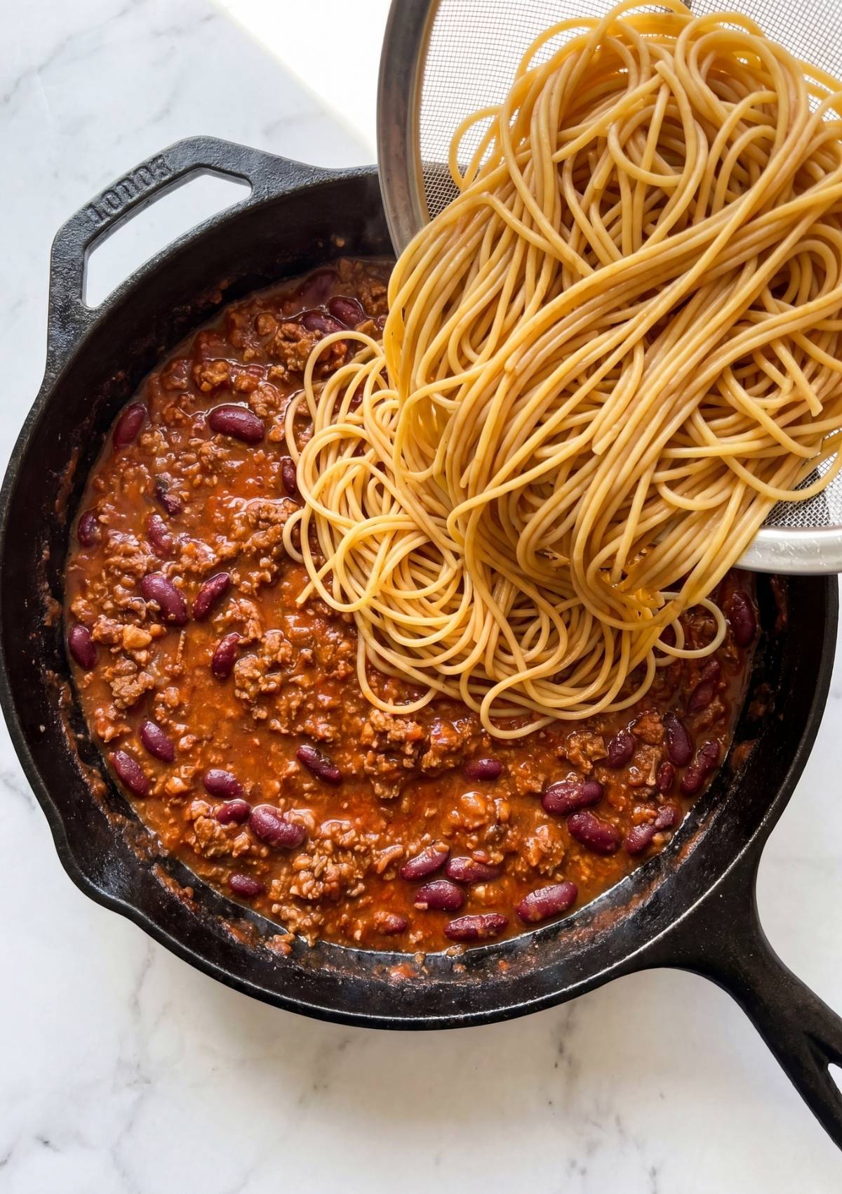 Cooked spaghetti noodles being added to a skillet filled with a thick, beefy chili sauce and beans.