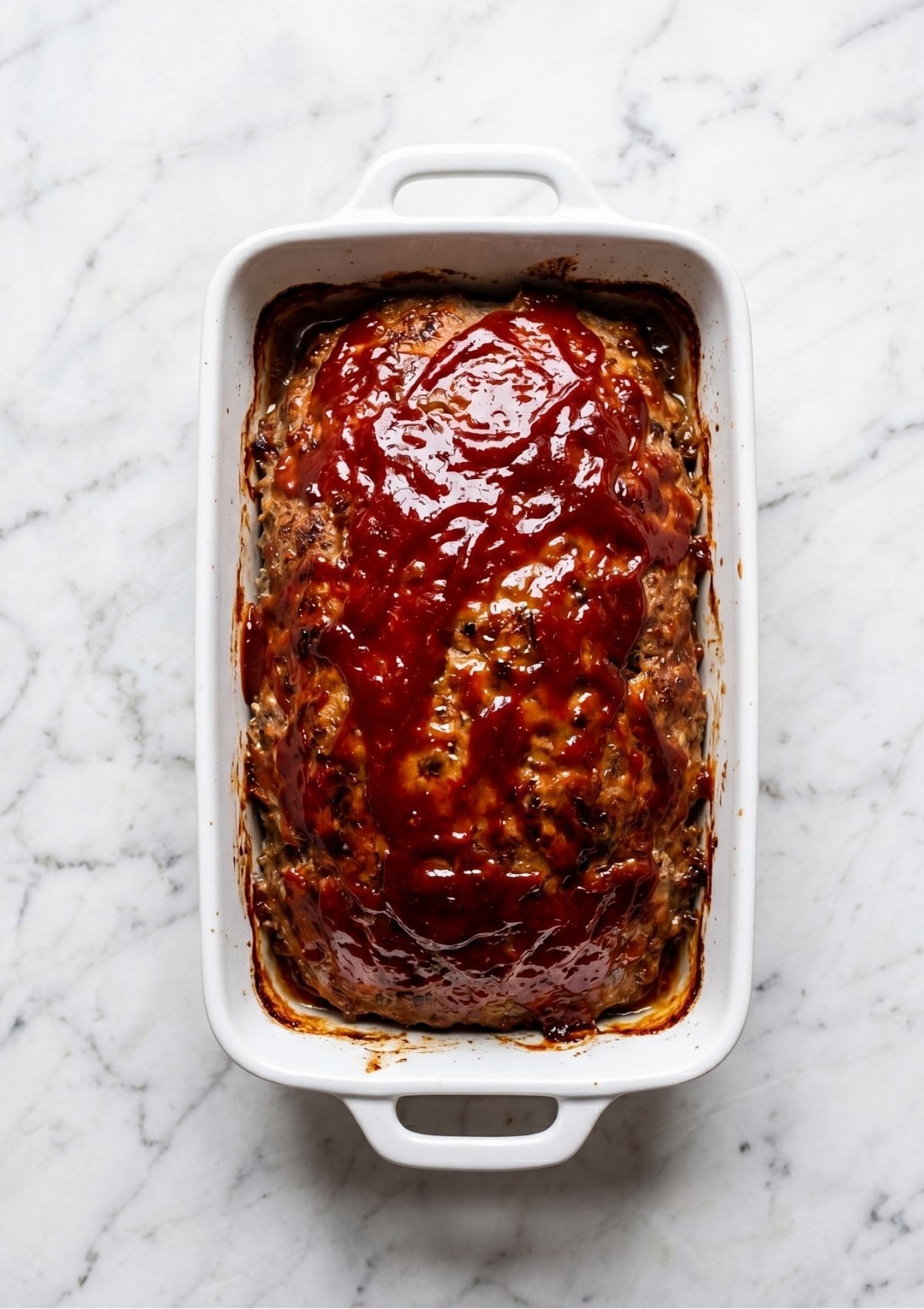 Baked meatloaf in a white rectangular dish covered in a thick red Meatloaf Glaze Recipe.