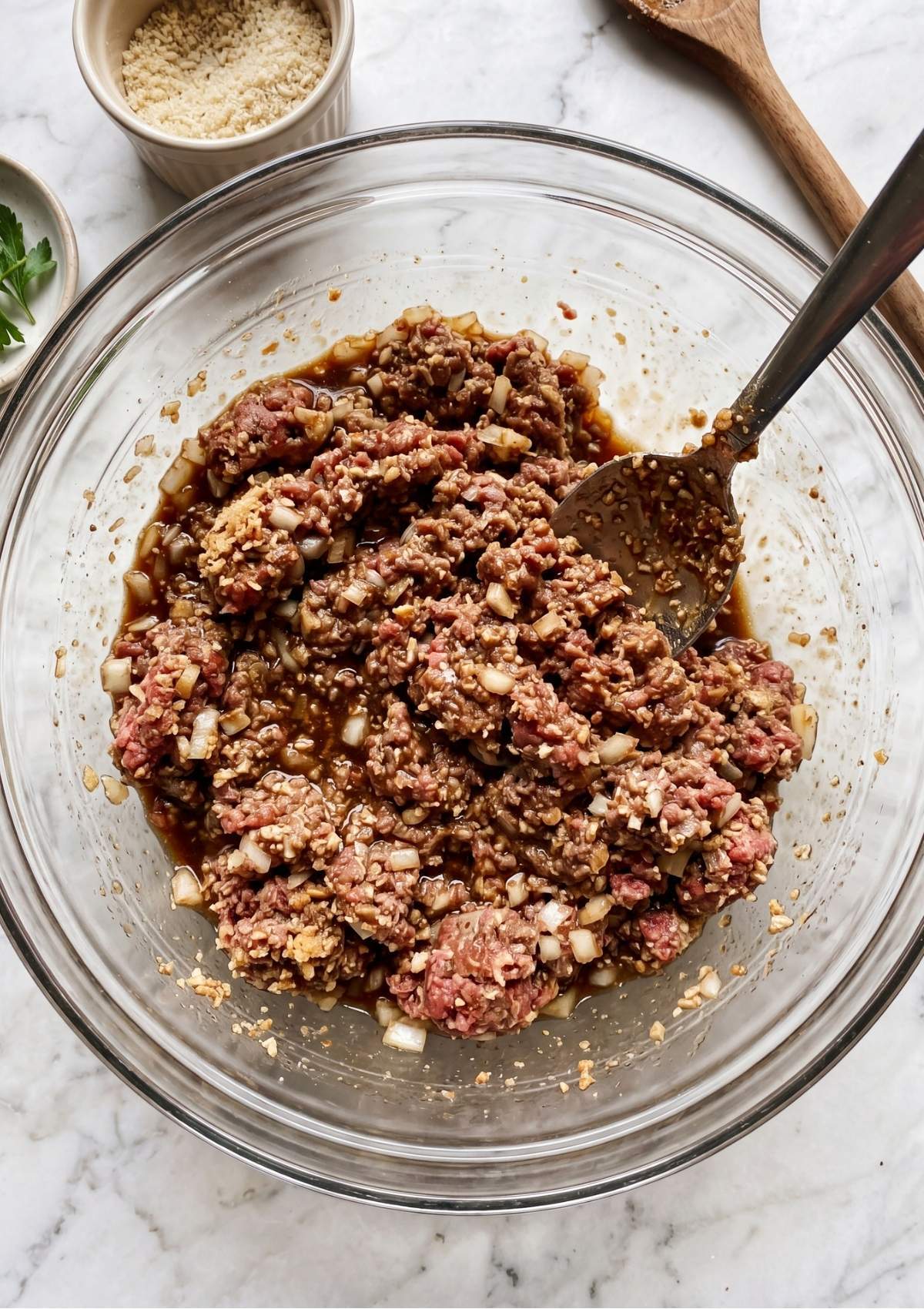 Ground beef being mixed with onions, breadcrumbs, and sauce in a glass bowl.