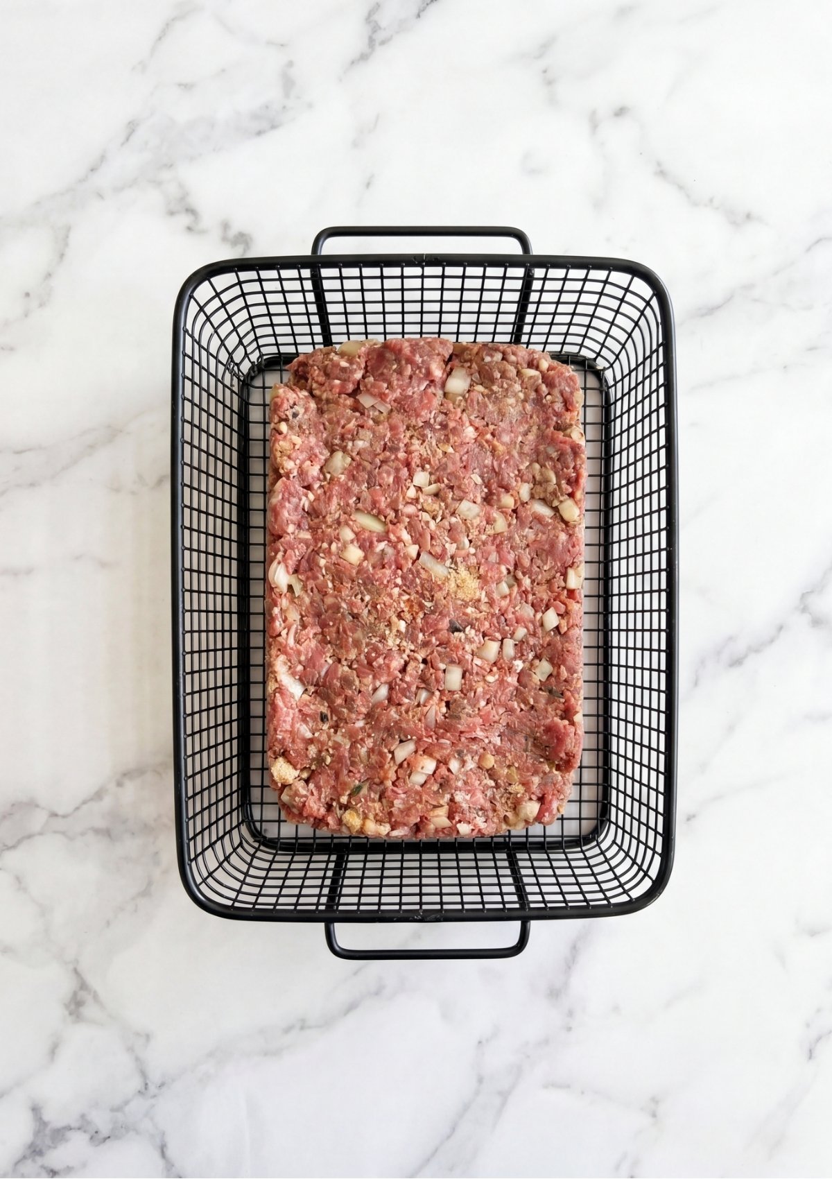 A rectangular base of raw ground beef mixture pressed into the bottom of a black wire air fryer or smoker basket on a white marble background.