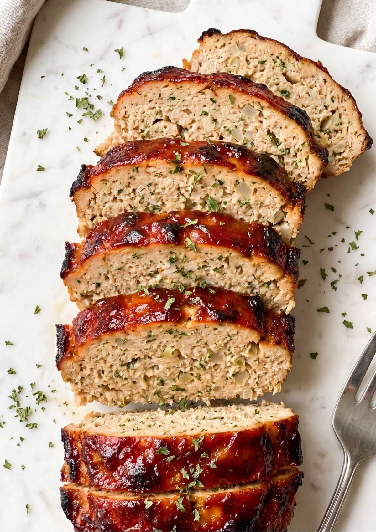 Thick slices of moist chicken meatloaf with visible herbs and onions arranged on a white marble surface.