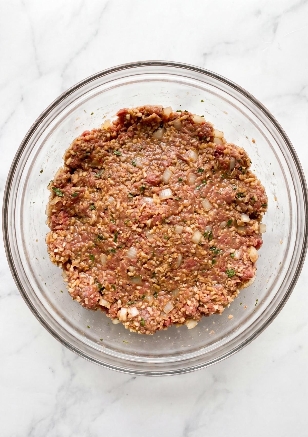 A glass mixing bowl on a marble surface containing the combined meatloaf ingredients including ground beef, herbs, and onions.