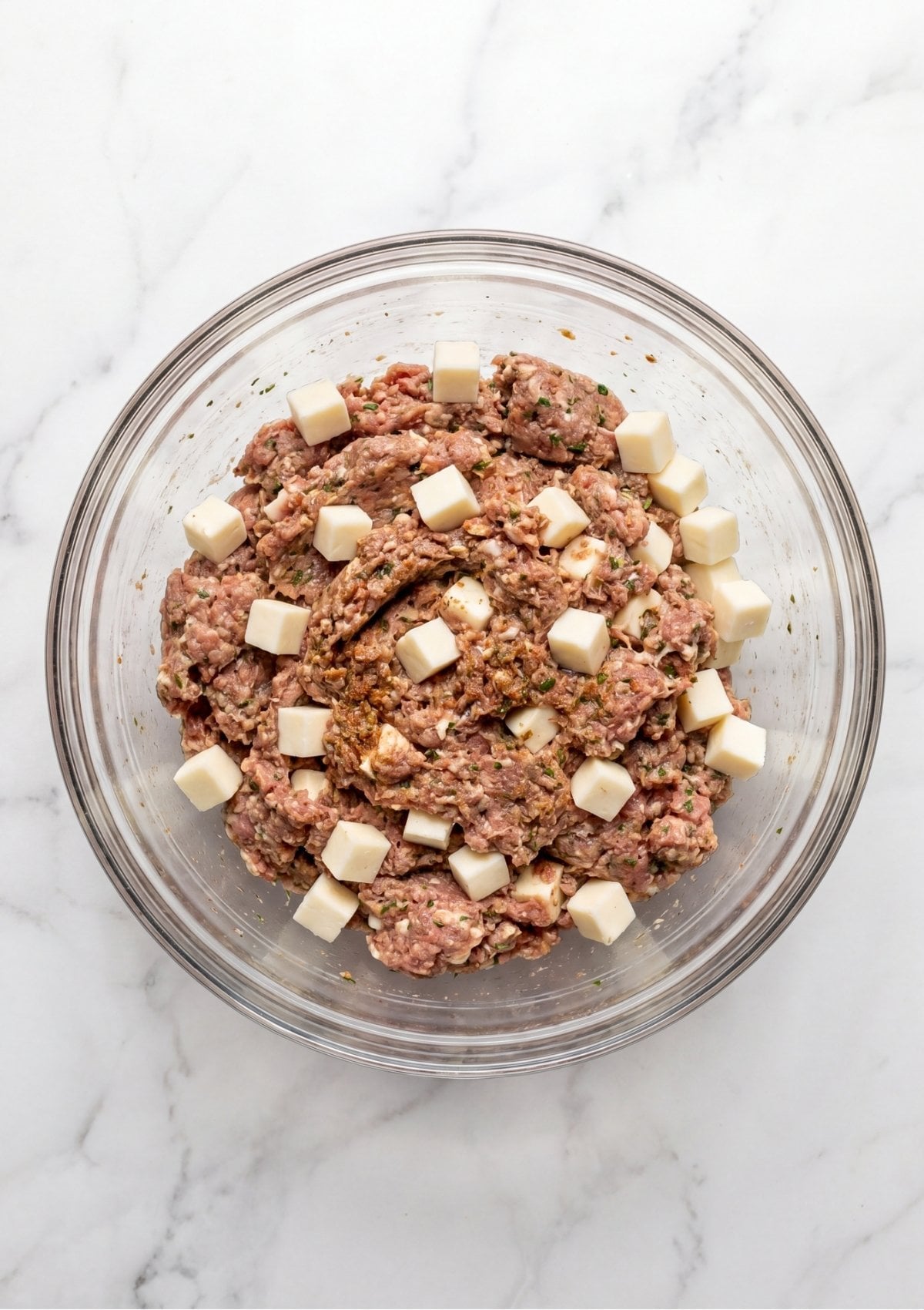 Raw meatloaf mixture in a glass bowl with large cubes of mozzarella cheese being folded in.