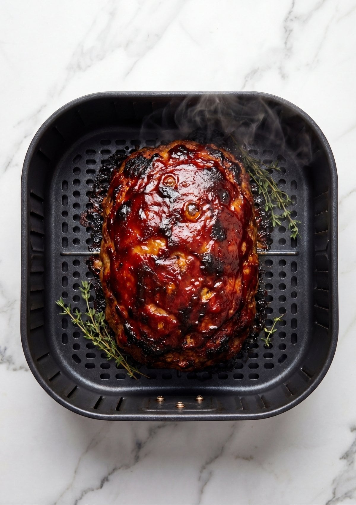 A glazed meatloaf cooking inside a black air fryer basket with fresh thyme sprigs on the side.