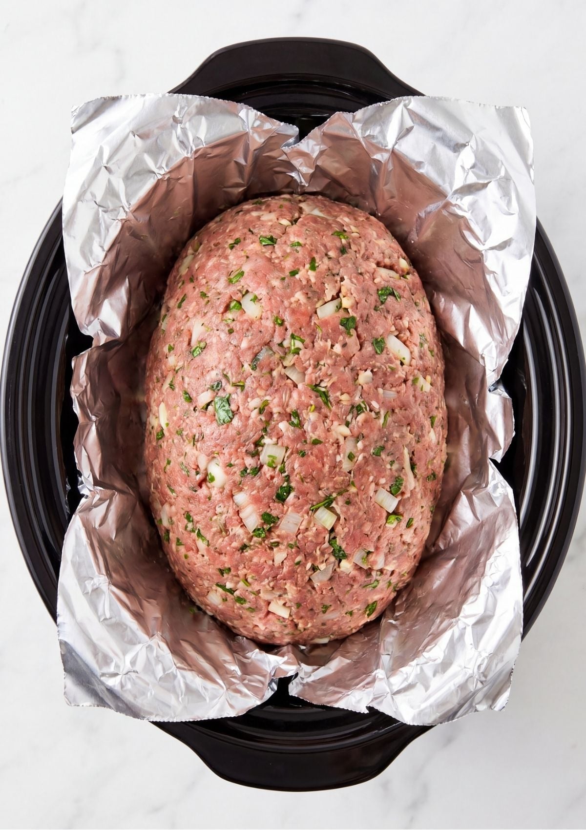 Raw meatloaf shaped into an oval and placed inside a slow cooker lined with silver foil
