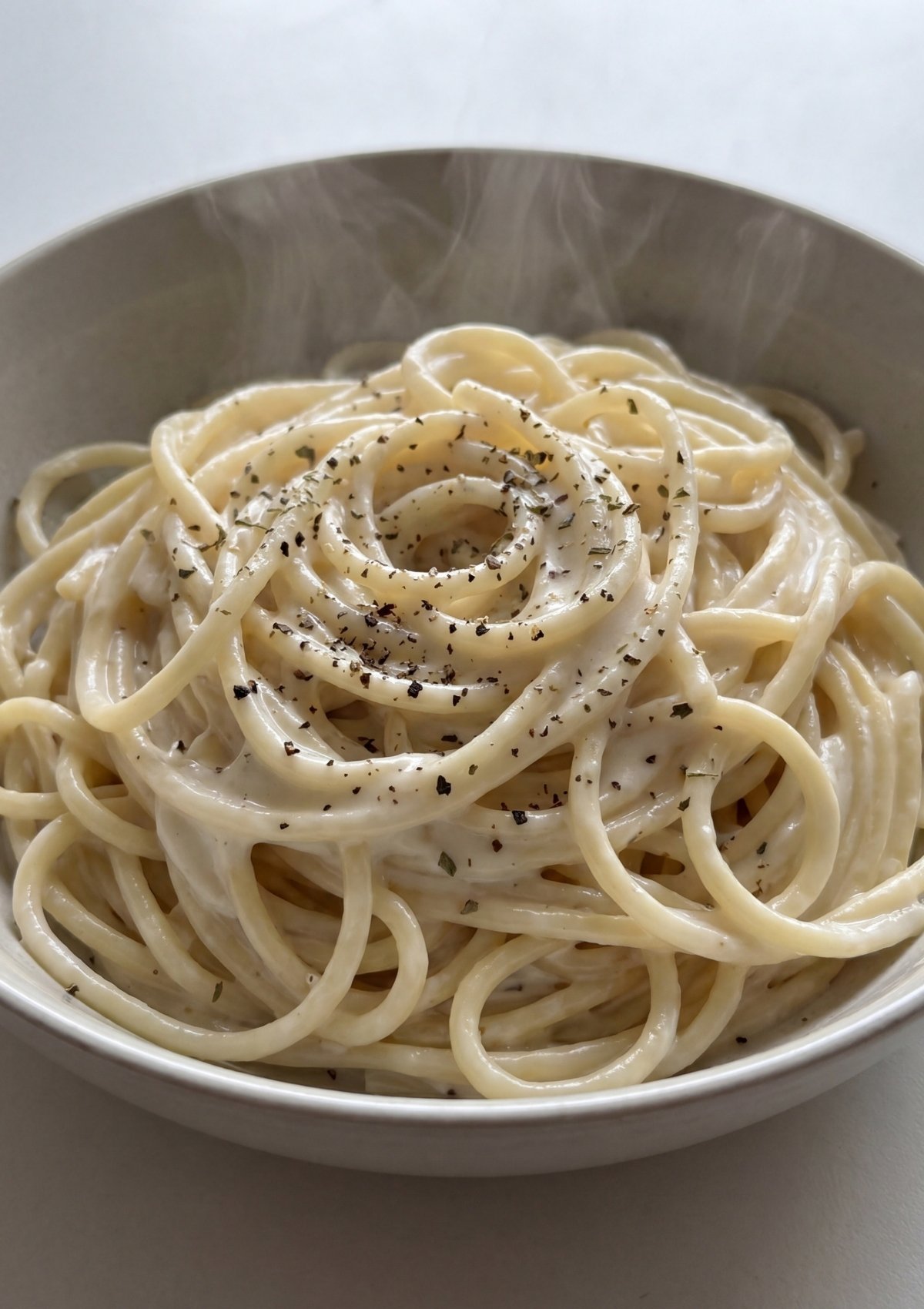 A white bowl containing steaming hot spaghetti tossed in a smooth, thick cream cheese sauce and garnished with cracked black pepper and dried herbs.