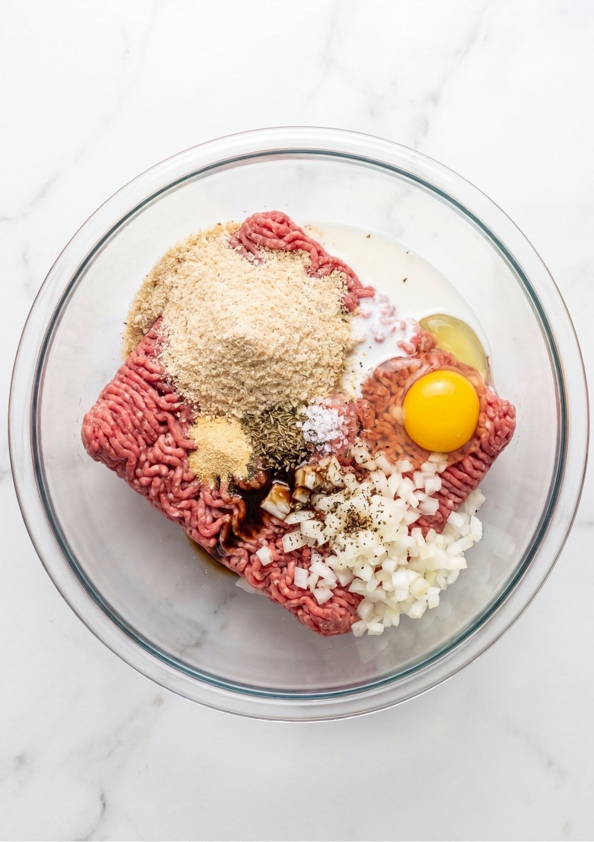 Clear glass mixing bowl containing raw ground beef, breadcrumbs, a whole egg, milk, and diced onions.