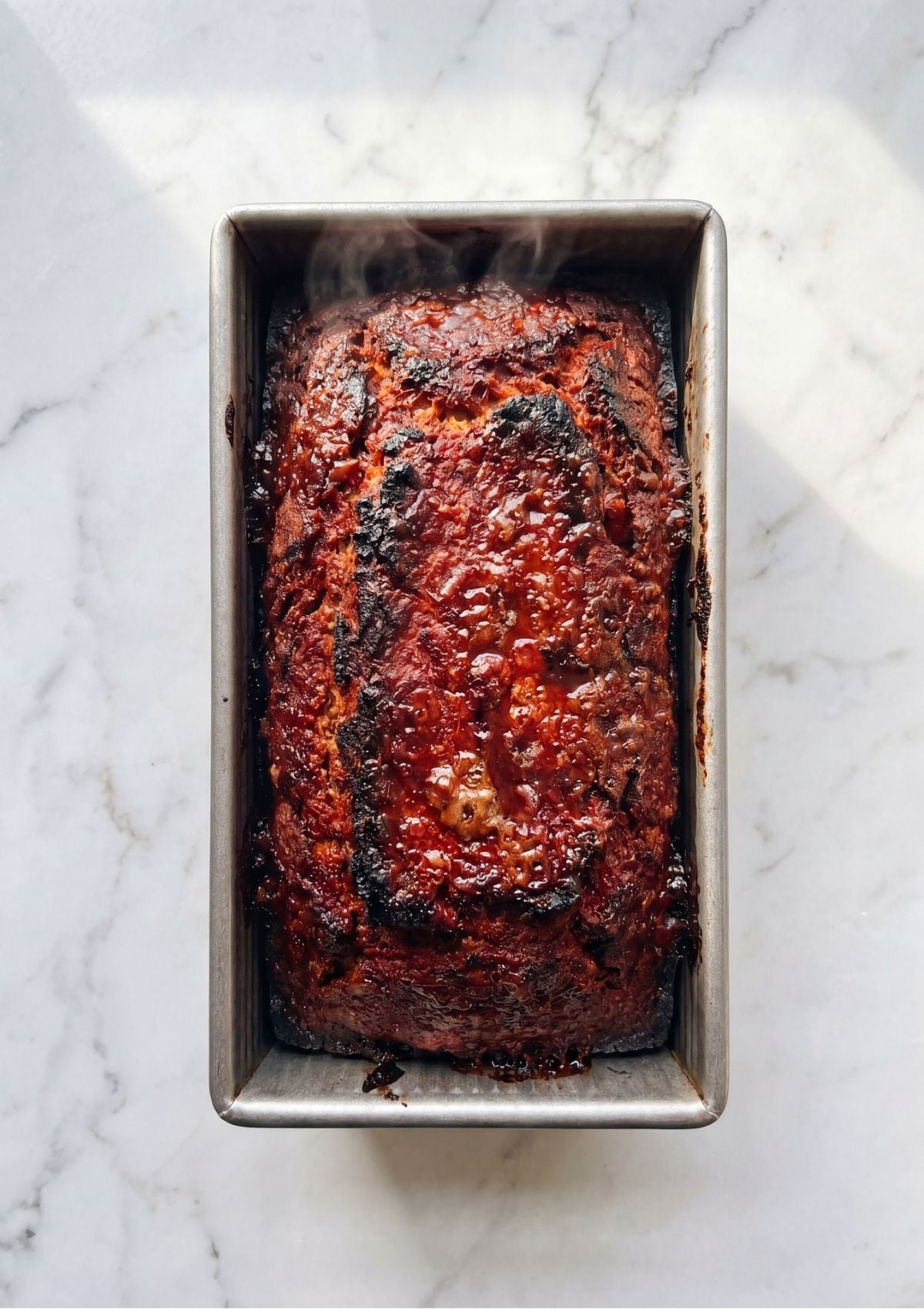 A top down view of a fully baked vegan meatloaf with a dark caramelized red glaze in a metal pan on a marble surface