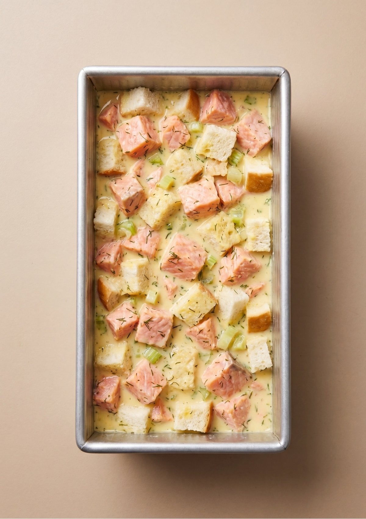 Cubed bread, fresh dill, and chunks of salmon mixed with an egg and milk batter inside a metal loaf pan before baking