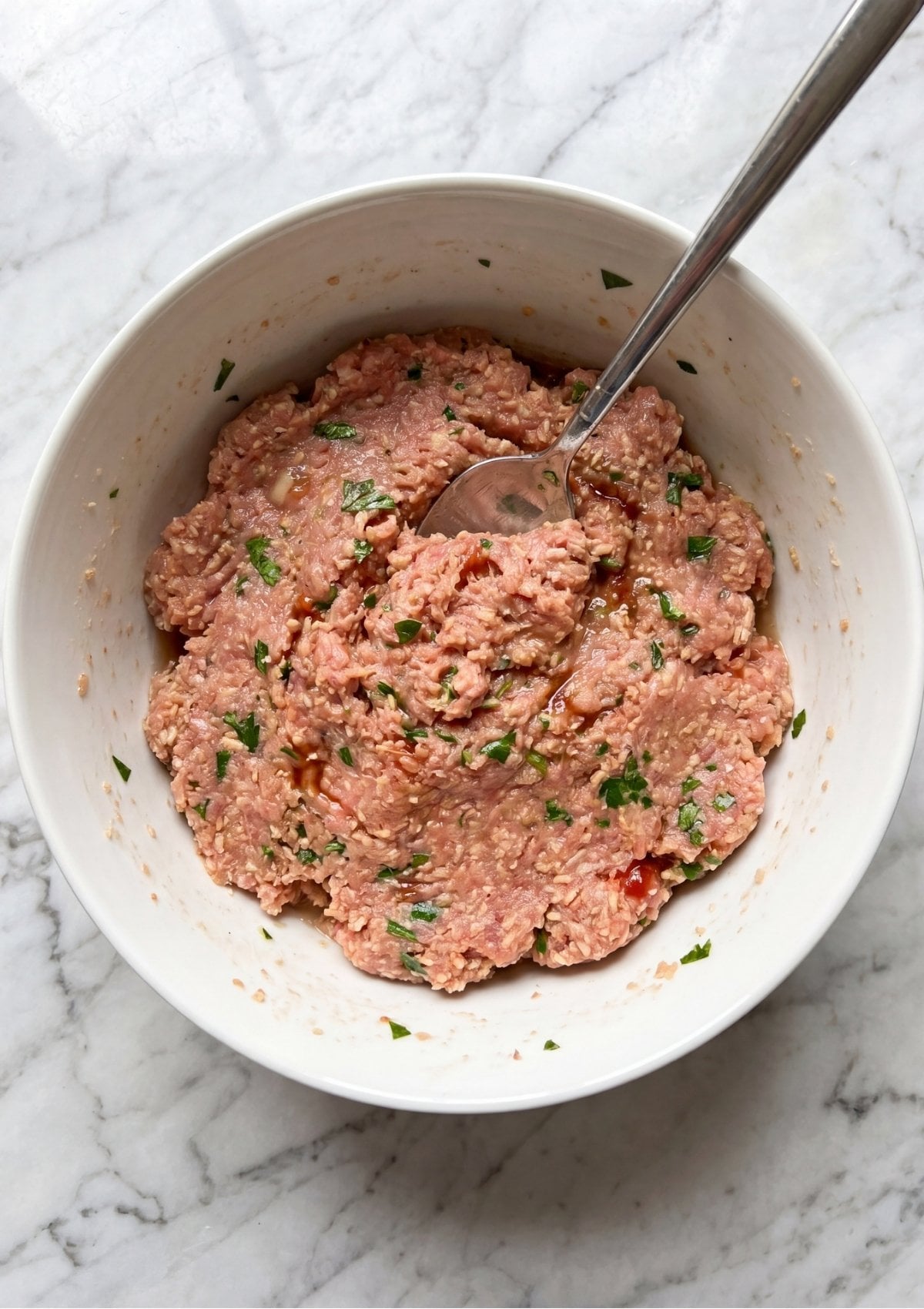 Ground turkey meatloaf mixture with fresh green parsley and spices being stirred in a white ceramic bowl with a silver spoon.