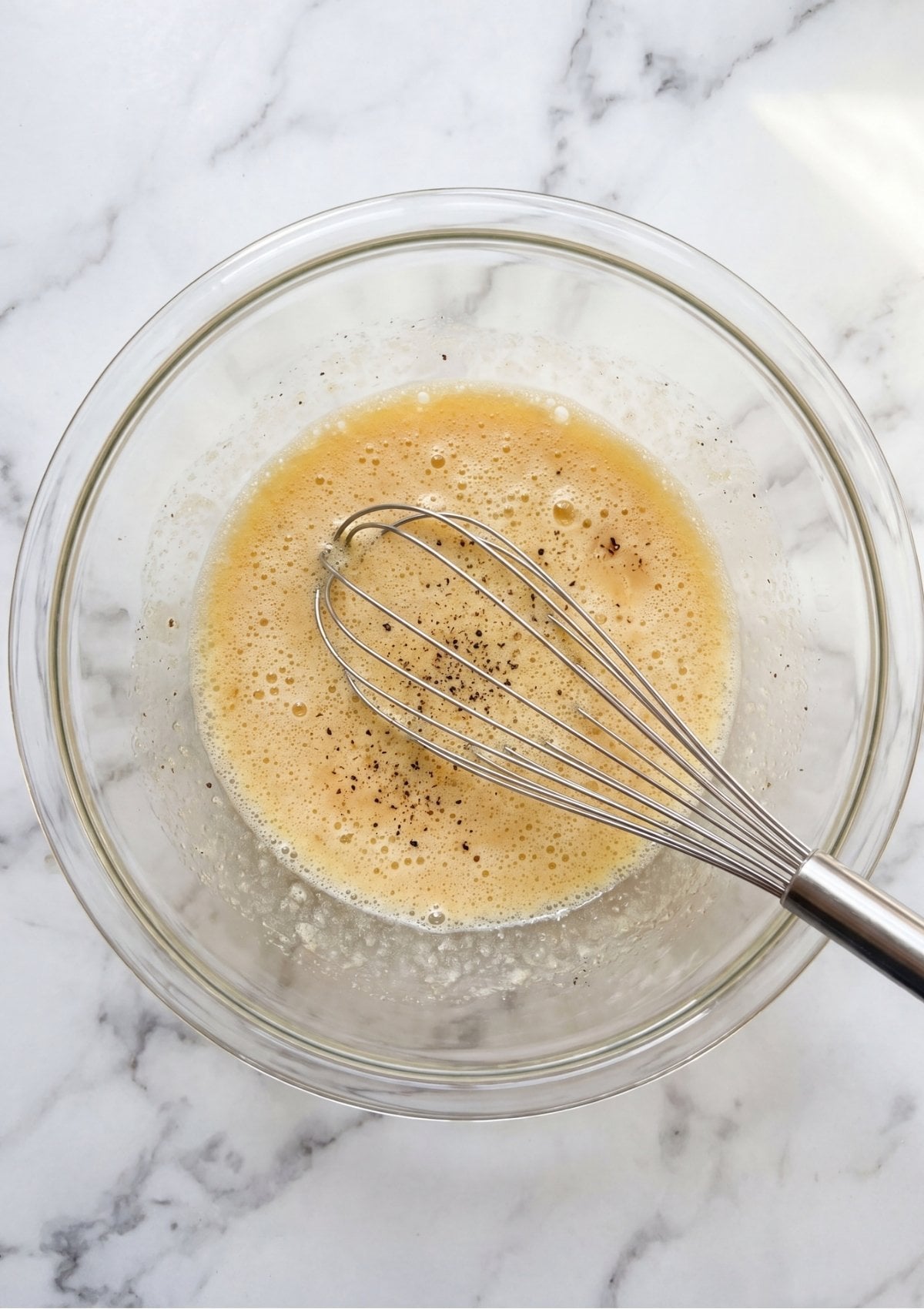 A stainless steel whisk blending eggs, milk, and spices into a frothy mixture inside a glass mixing bowl.