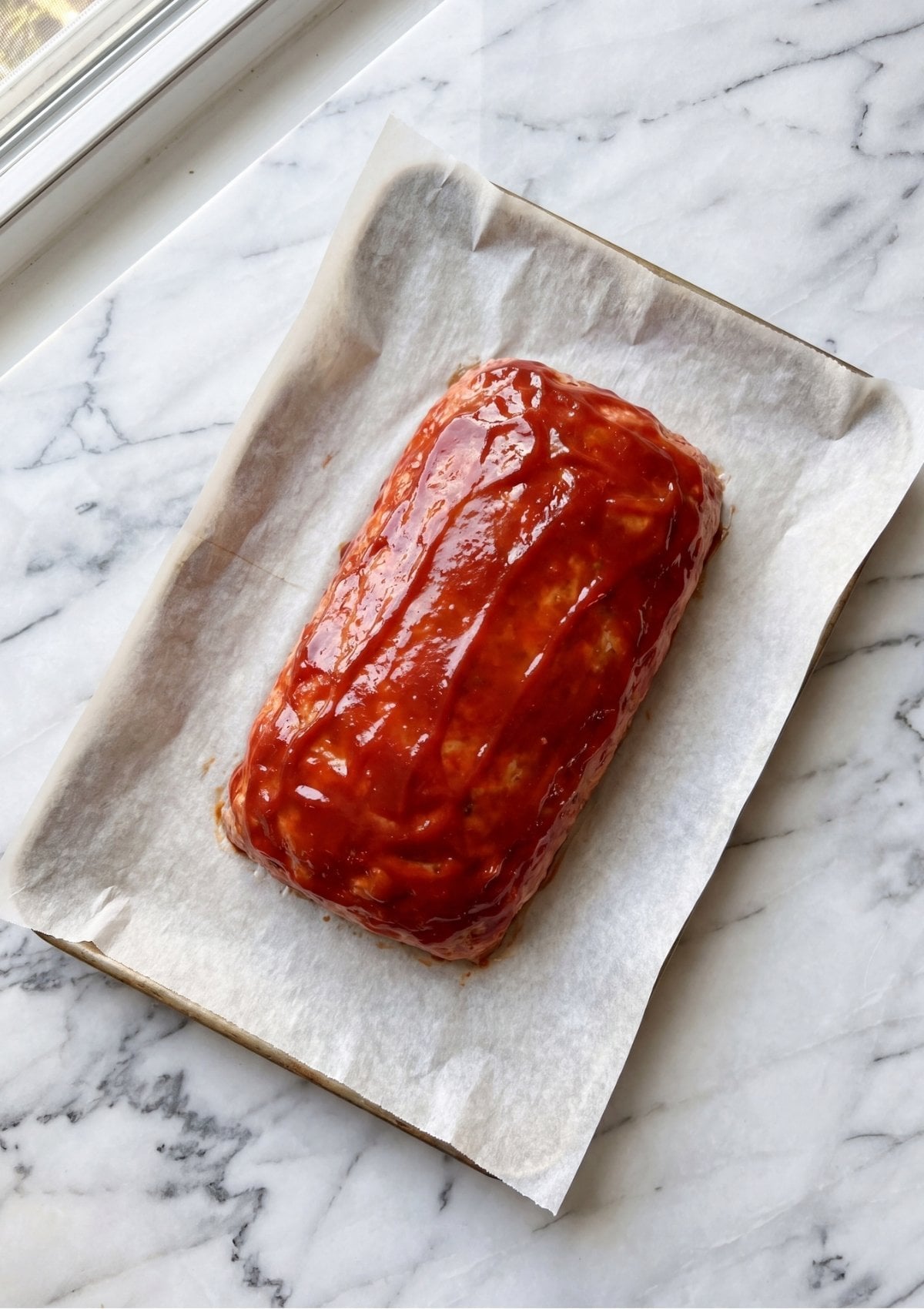 An unbaked, shaped turkey meatloaf topped with smooth red ketchup glaze on a parchment-lined baking sheet
