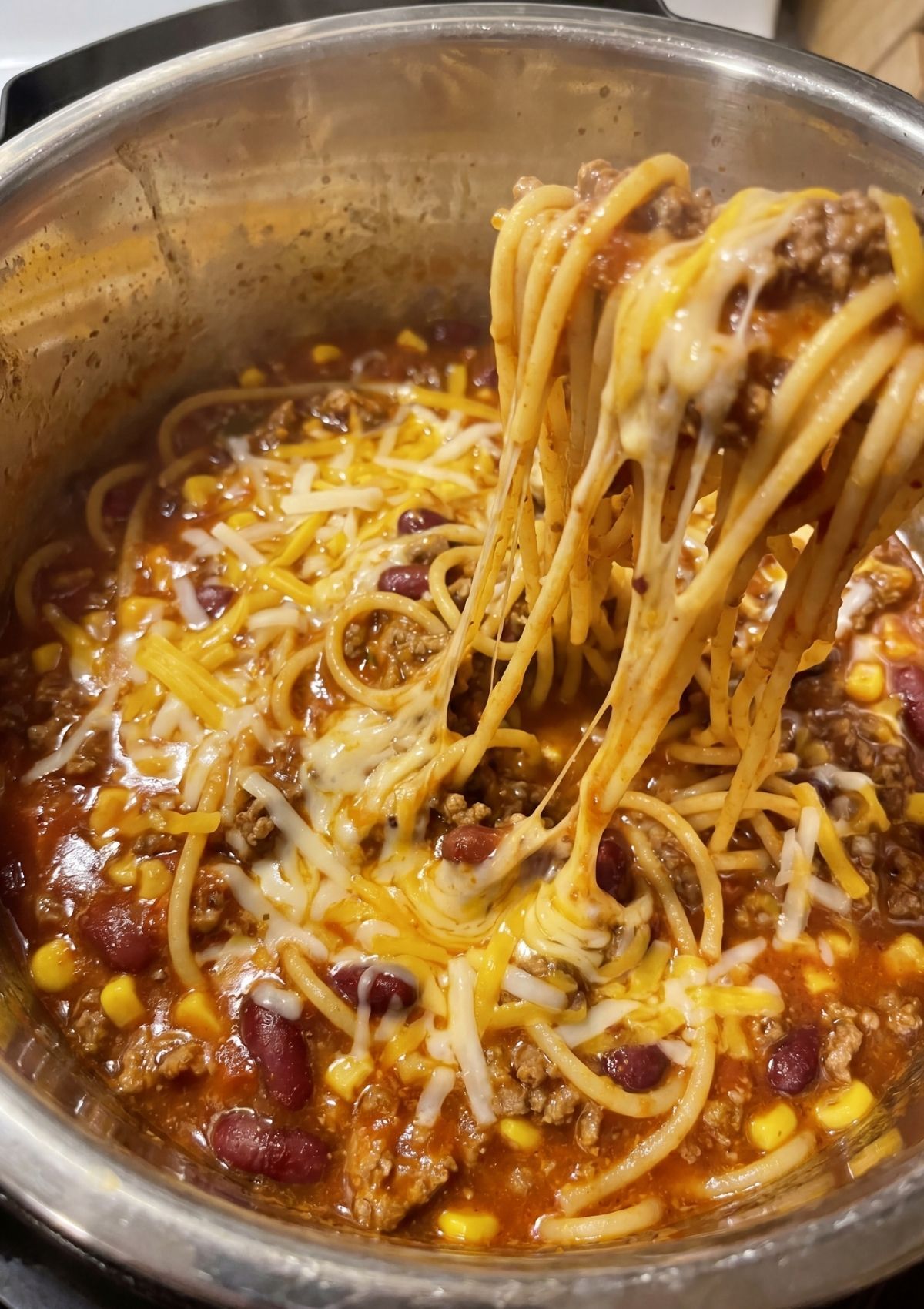 A wooden spoon lifting a serving of Instant Pot Cowboy Spaghetti from the pot, showing long strings of melted cheese, corn, and kidney beans.