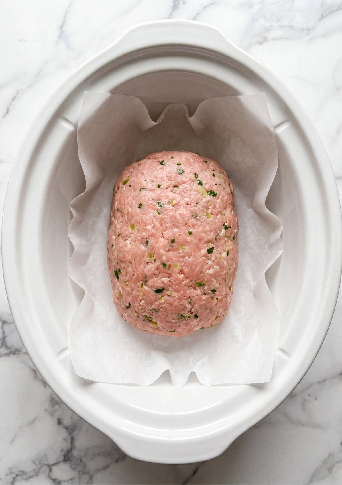 Raw seasoned ground turkey mixed with fresh green herbs shaped into a loaf inside a slow cooker lined with parchment paper