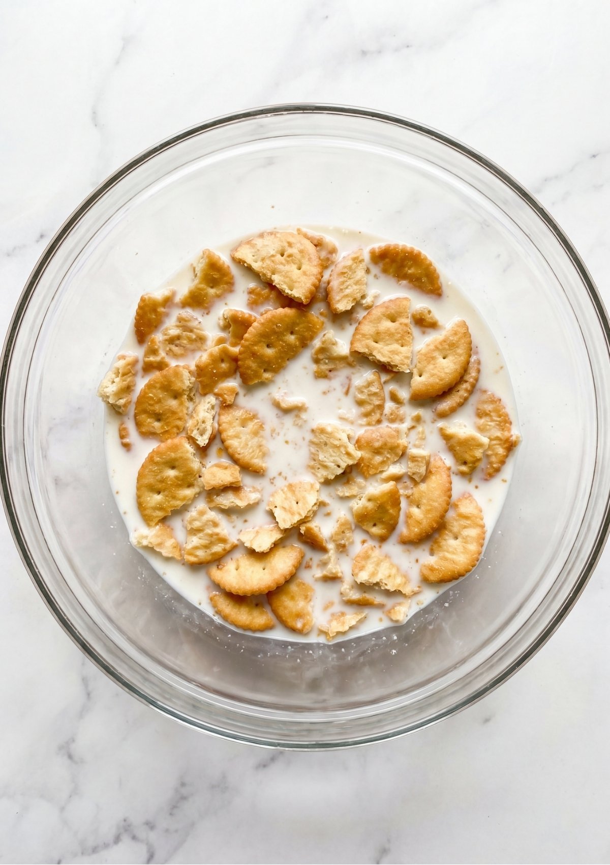 Crushed buttery round crackers soaking in a clear glass bowl of milk on a marble countertop