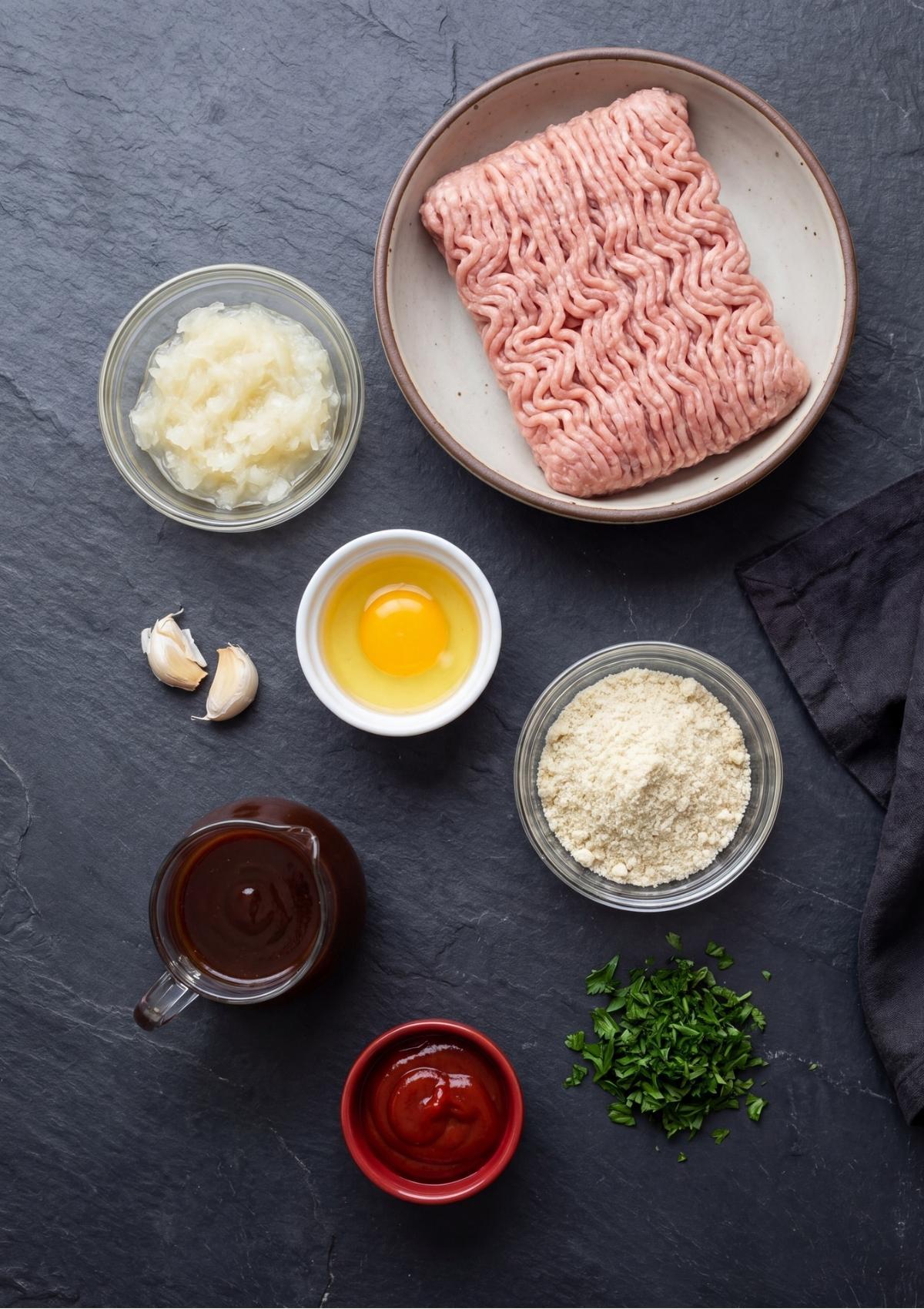 Overhead view of ingredients for turkey meatloaf including ground turkey, grated onion, a raw egg, almond flour, BBQ sauce, ketchup, and chopped parsley.
