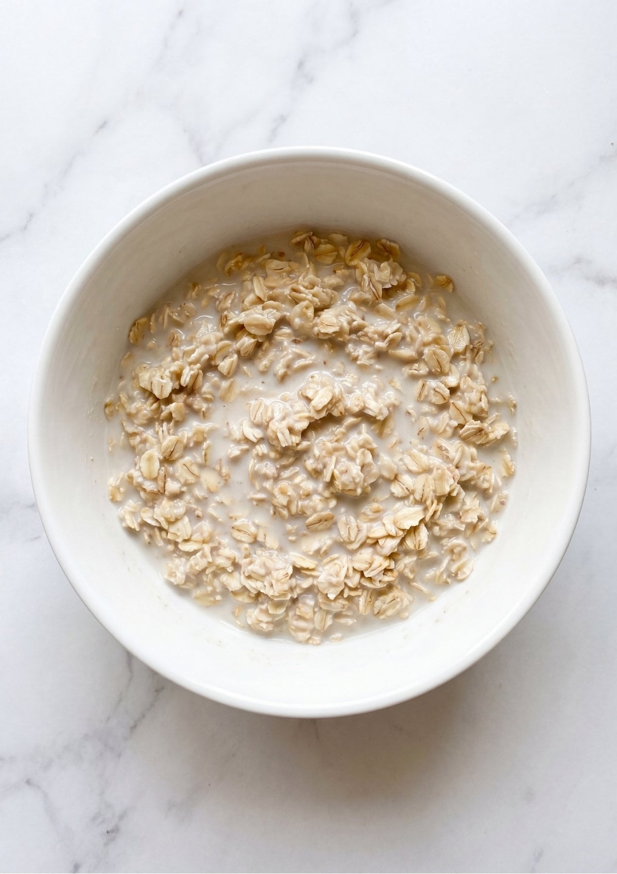 Rolled oats soaking in a pool of white milk inside a white ceramic mixing bowl.