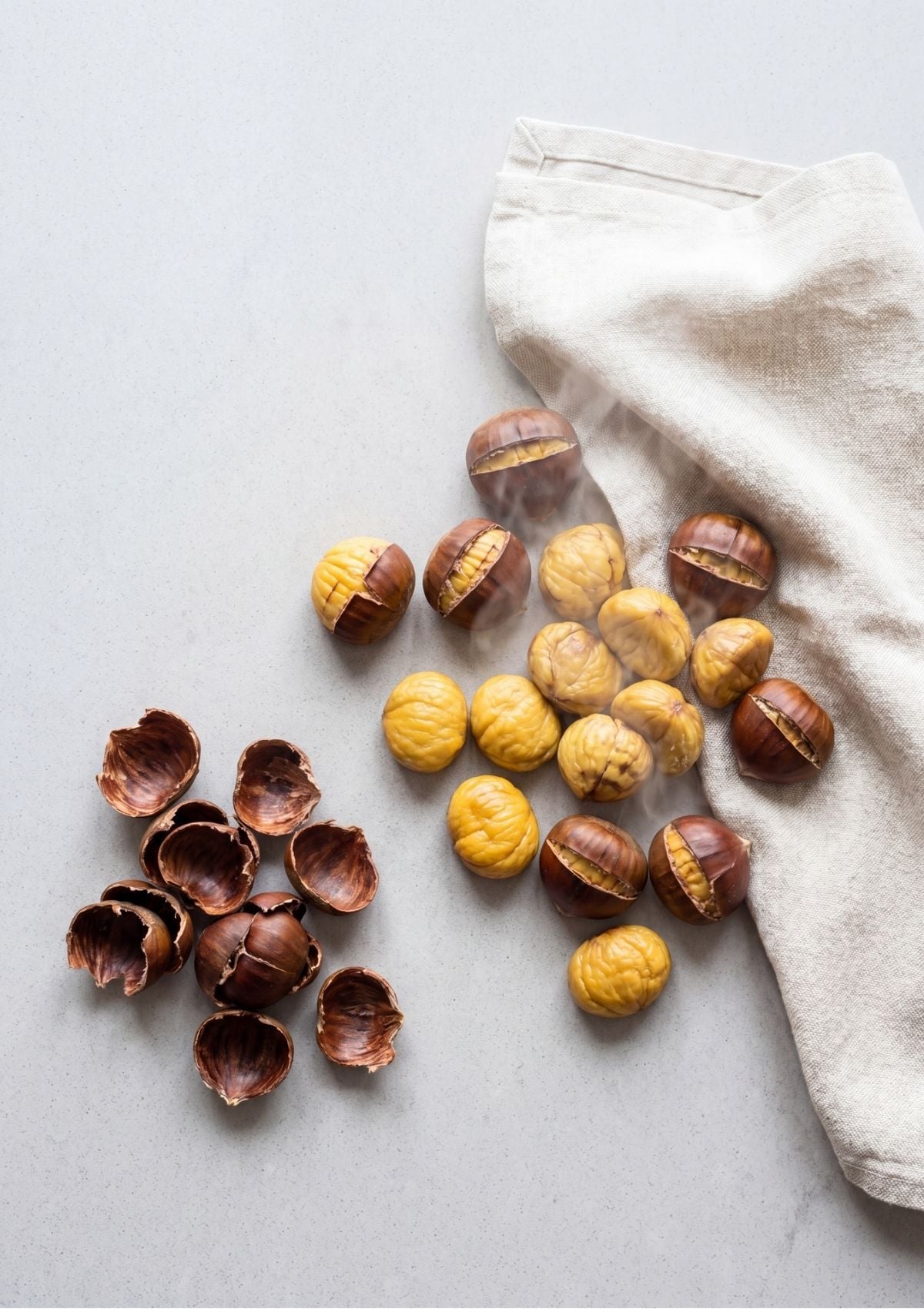 Steam rising from peeled golden chestnuts next to a pile of empty roasted brown shells on a light grey surface.