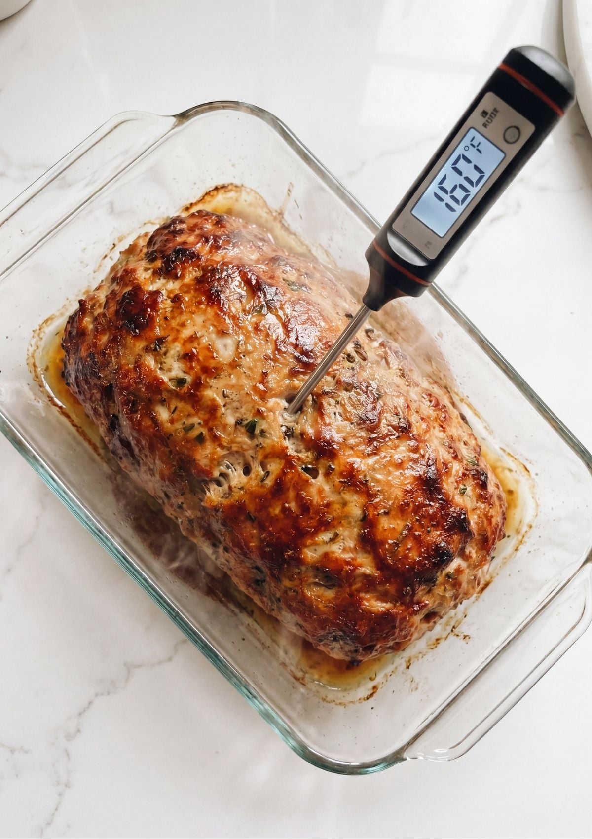 A digital meat thermometer inserted into a golden brown baked meatloaf inside a glass baking dish showing 160 degrees