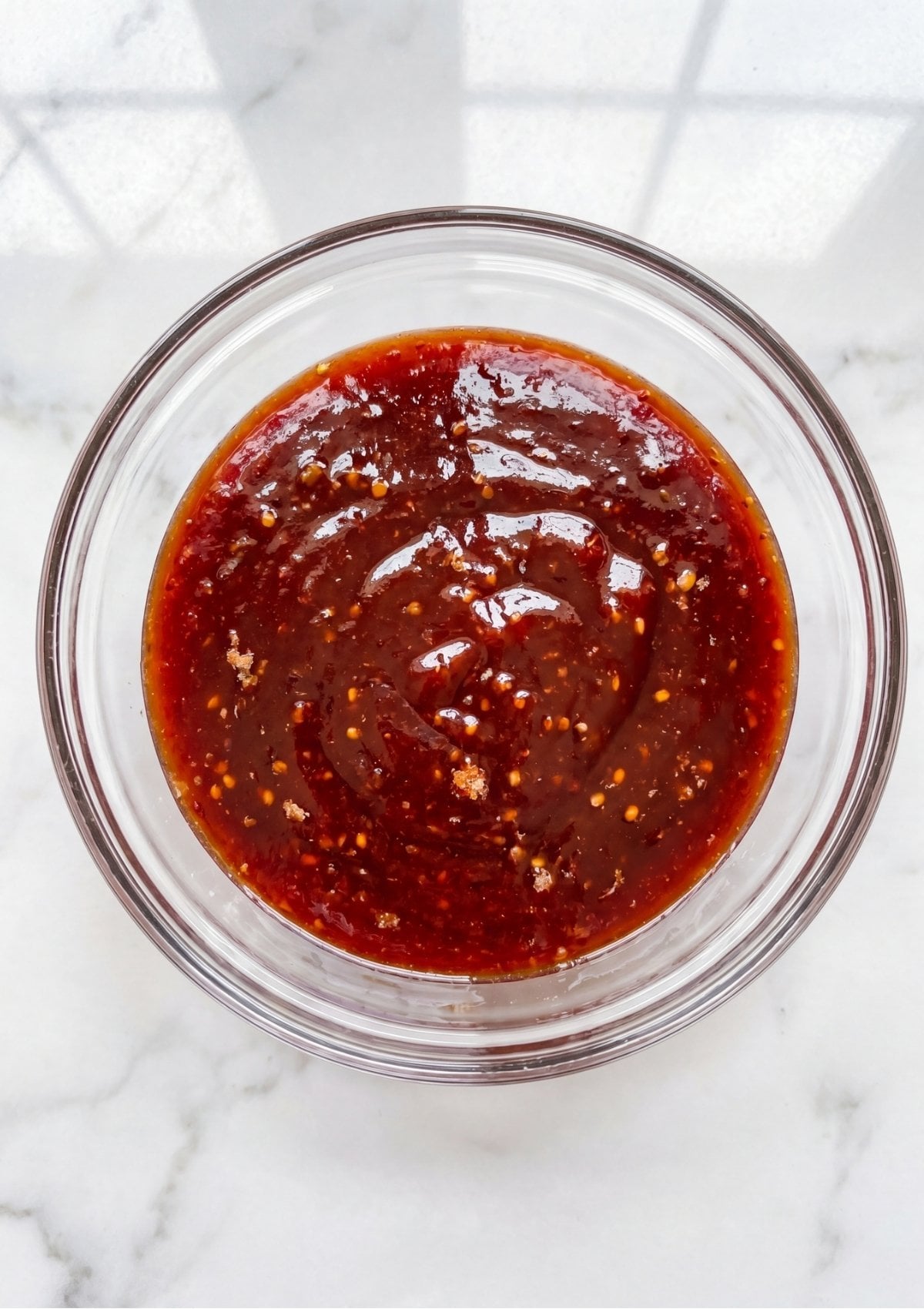 A glass bowl filled with a dark red meatloaf glaze sitting on a white marble countertop.