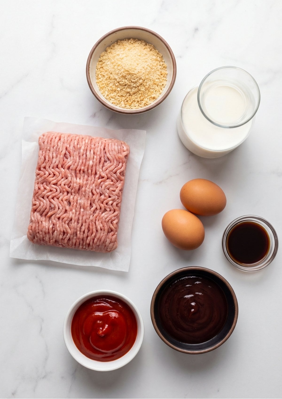 Flat lay of recipe ingredients including ground turkey, eggs, breadcrumbs, milk, ketchup, and savory sauces on a white marble surface.