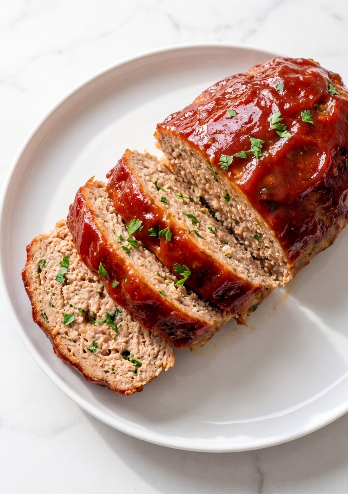 A sliced loaf of ground turkey meatloaf with a thick red glaze and fresh parsley garnish on a white ceramic plate.