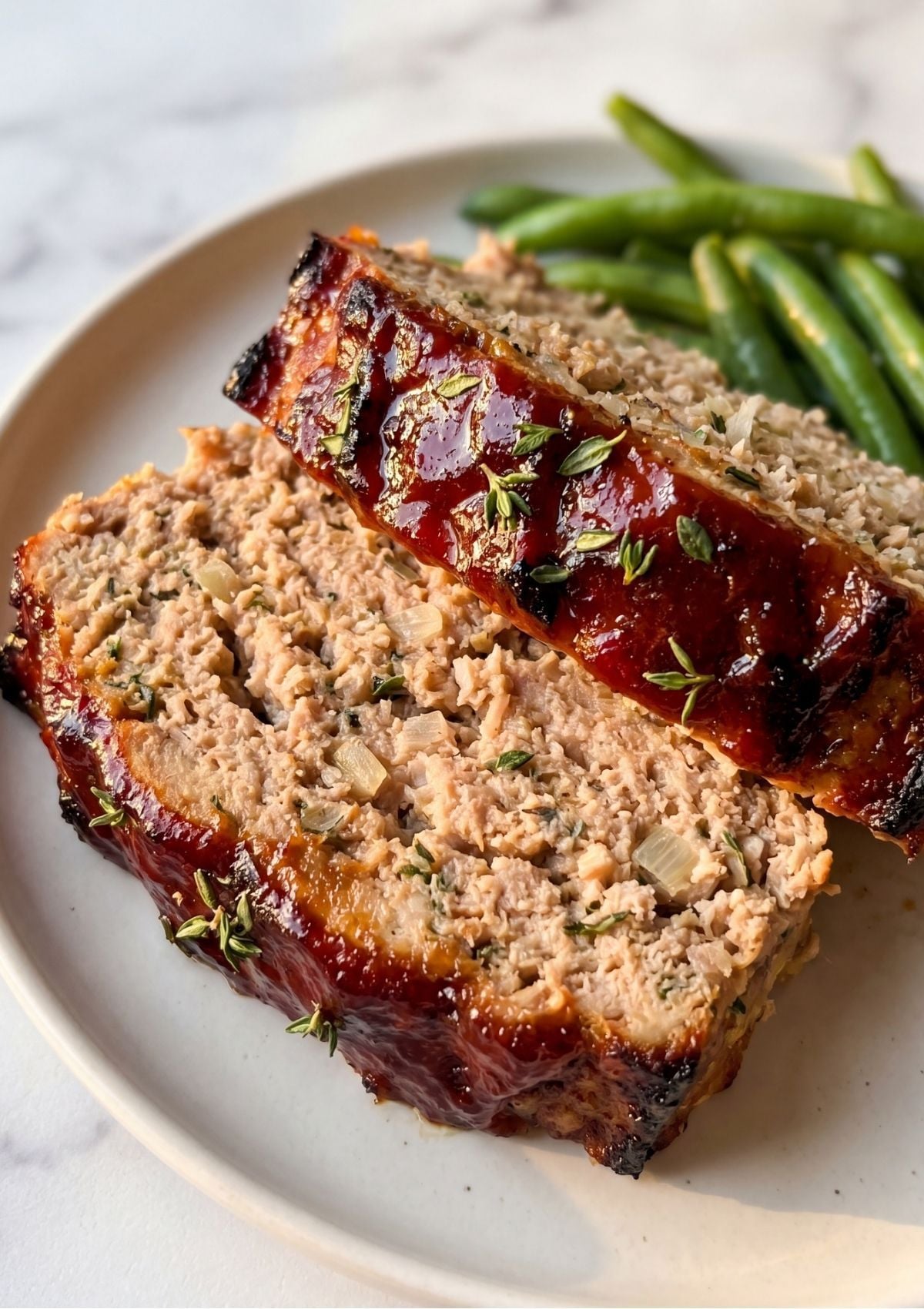 Two thick slices of juicy turkey meatloaf topped with a shiny red glaze and fresh herbs on a white plate.