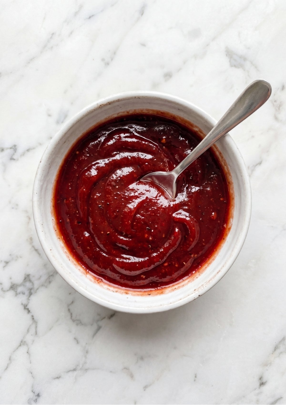 A small white bowl filled with a thick red ketchup-based meatloaf glaze and a silver spoon.