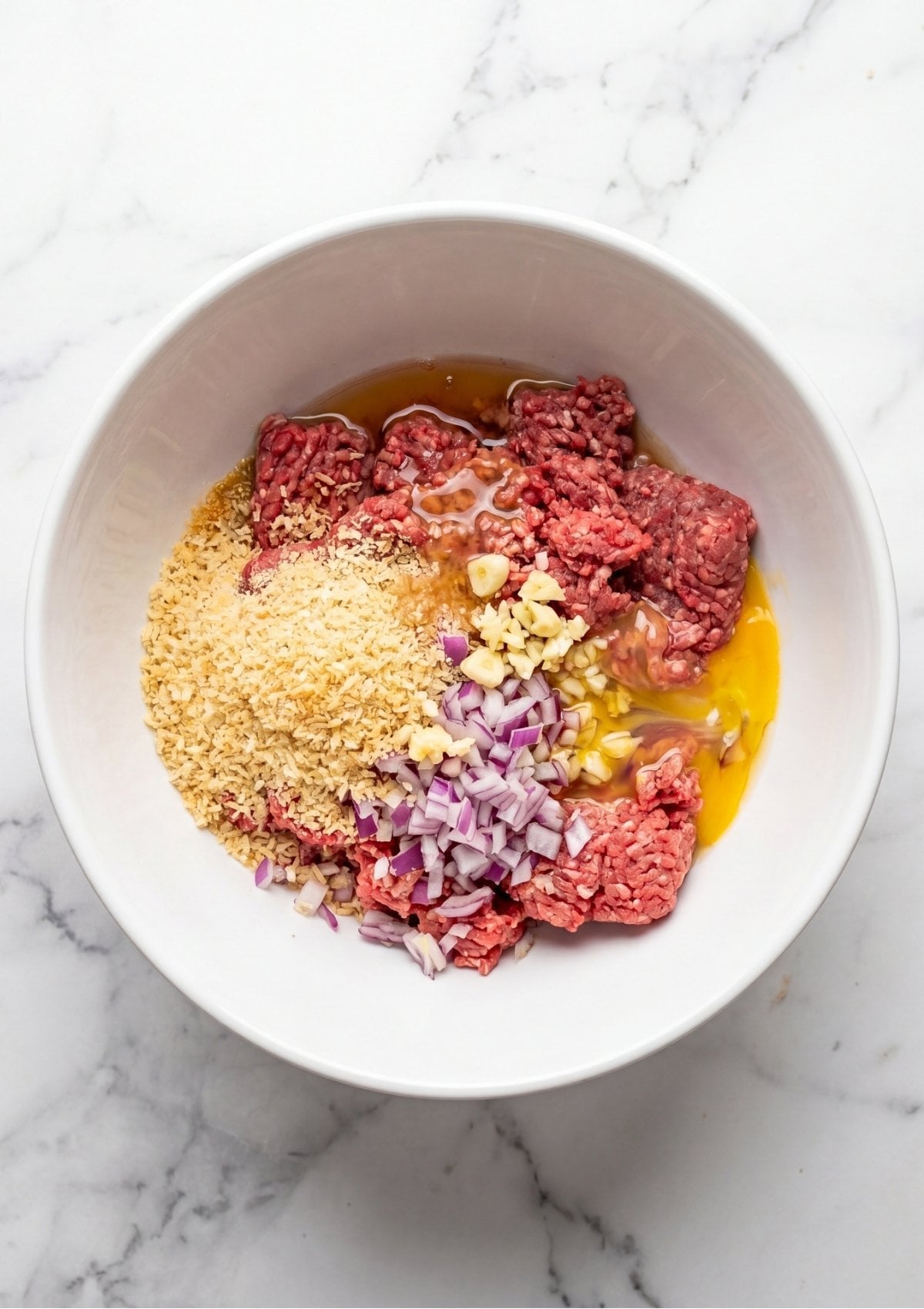 A top-down view of a white ceramic bowl containing ground beef topped with Panko breadcrumbs, minced garlic, diced red onions, and raw egg.