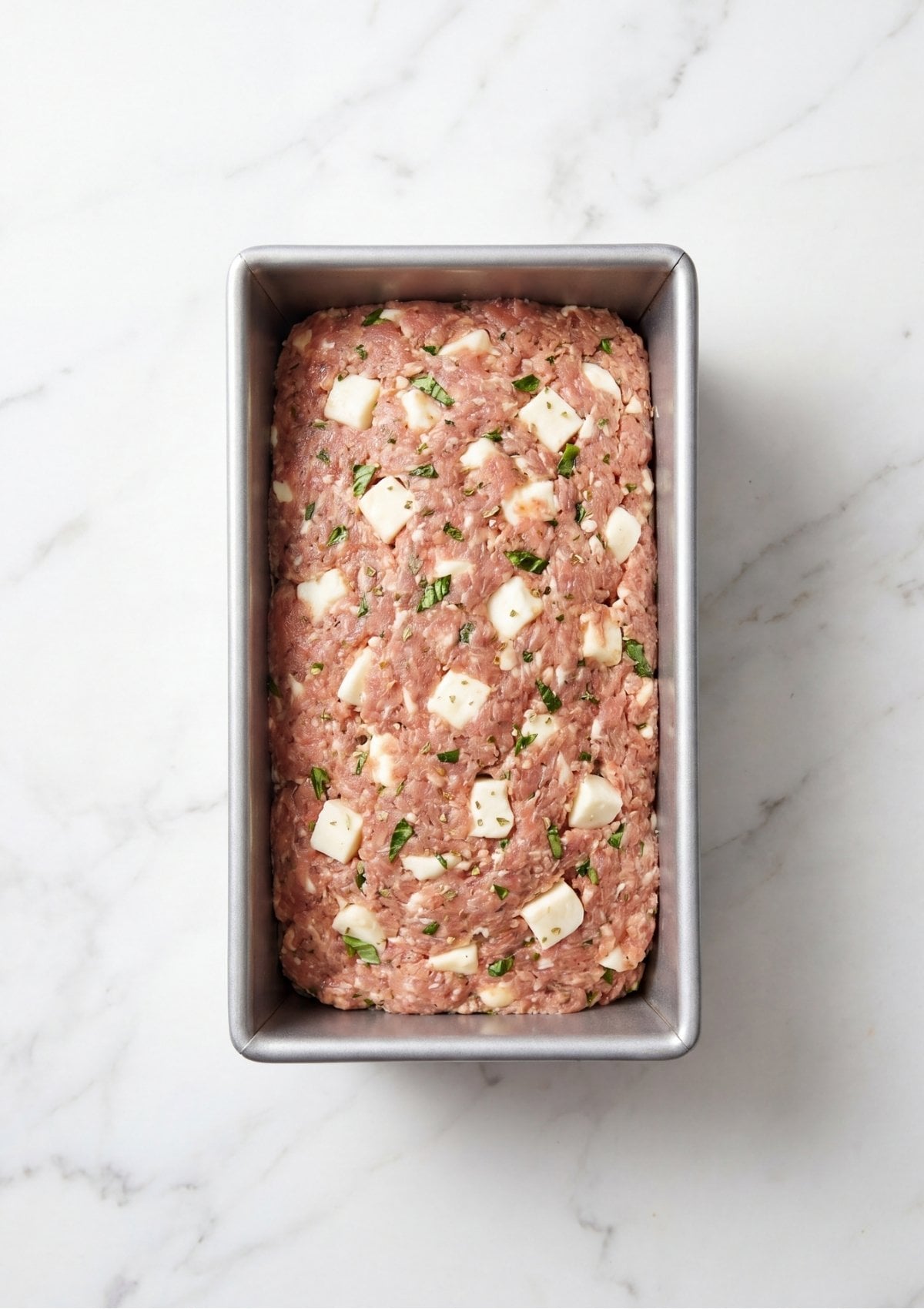 The raw meat and cheese mixture flattened evenly into a silver loaf pan before adding the topping.