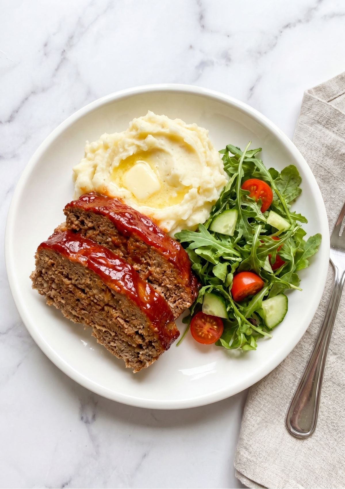 A top-down view of two thick slices of glazed meatloaf served on a white plate with a scoop of buttery mashed potatoes and a fresh arugula salad with cherry tomatoes and cucumber on a marble surface.