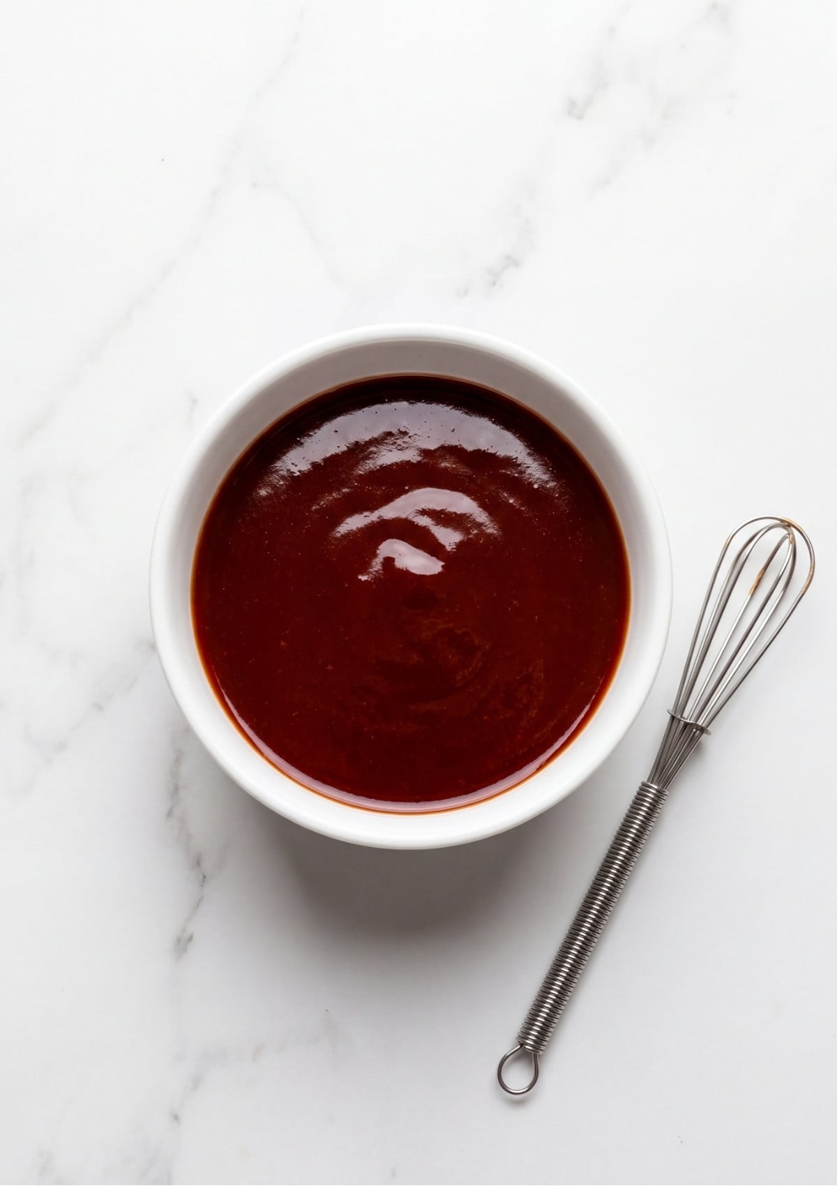 A small white bowl filled with a smooth dark red meatloaf glaze next to a wire whisk