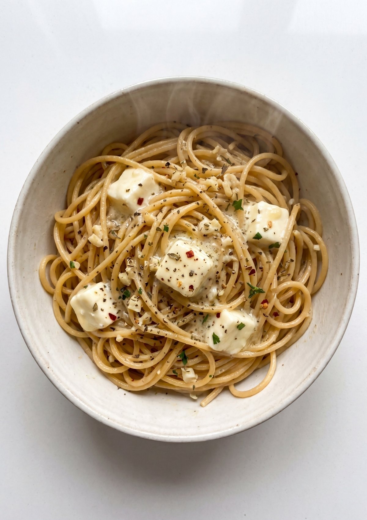 A top-down view of a white bowl where cubes of cream cheese, minced garlic, and red pepper flakes are being tossed with hot, cooked spaghetti.