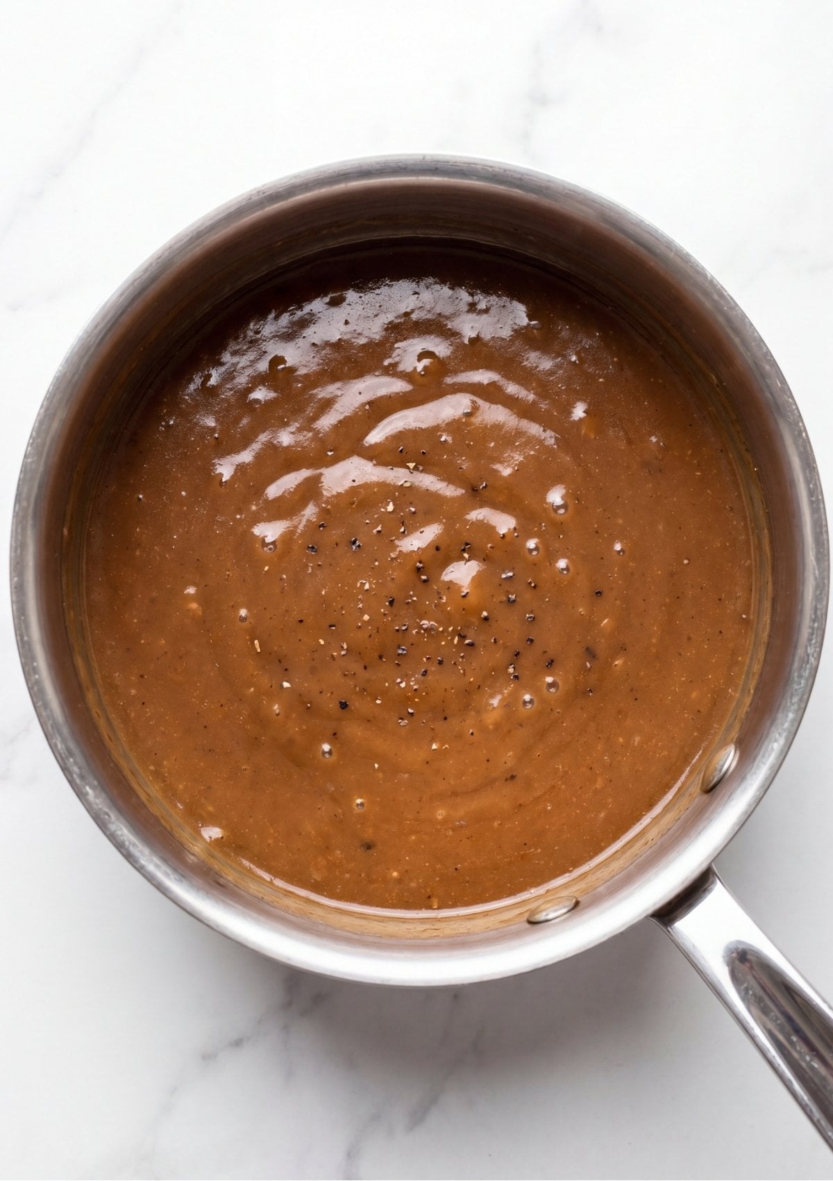 A stainless steel saucepan filled with smooth brown beef gravy seasoned with cracked black pepper