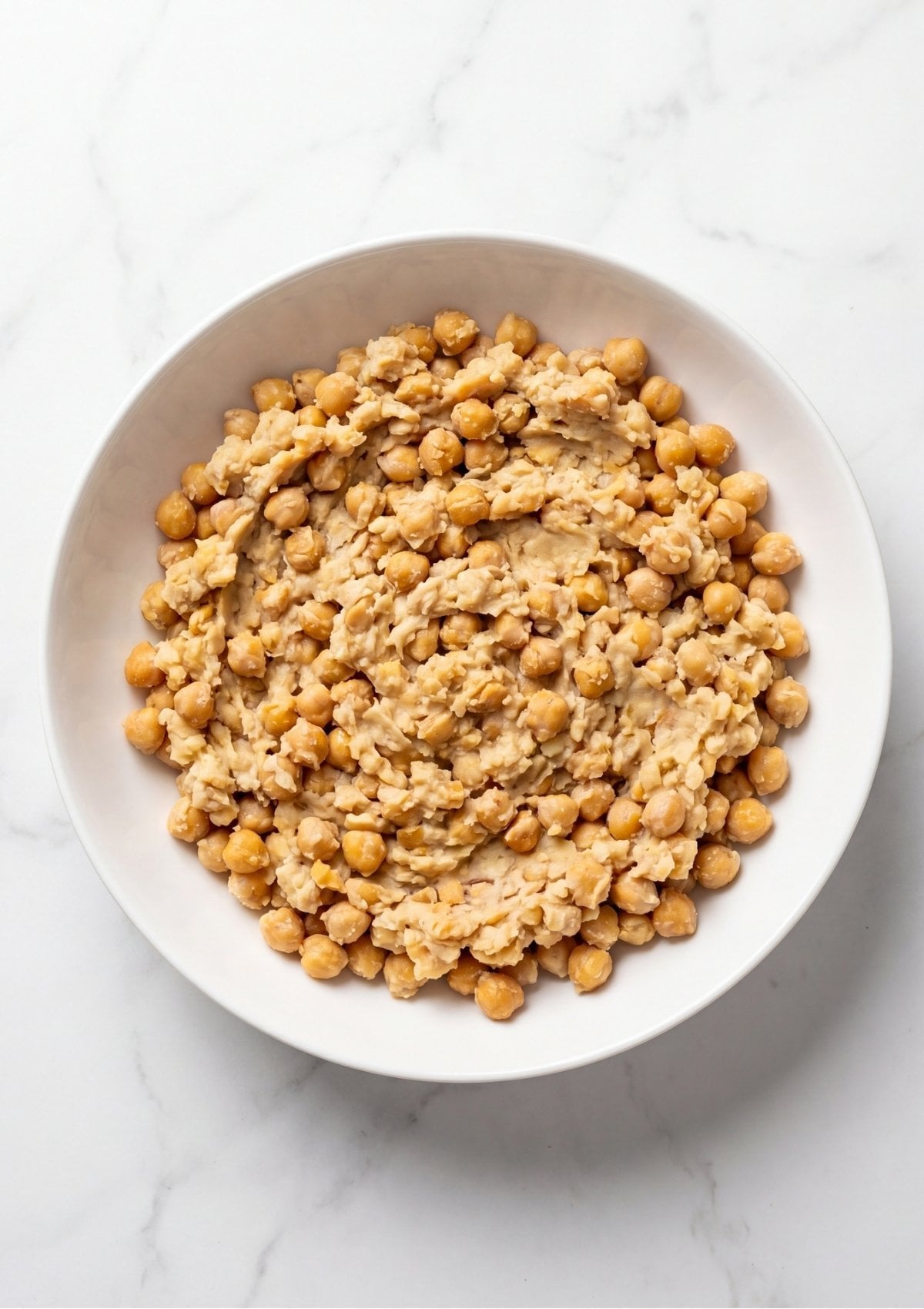 A white bowl filled with mashed chickpeas on a marble countertop showing a textured consistency for a vegan meatloaf recipe