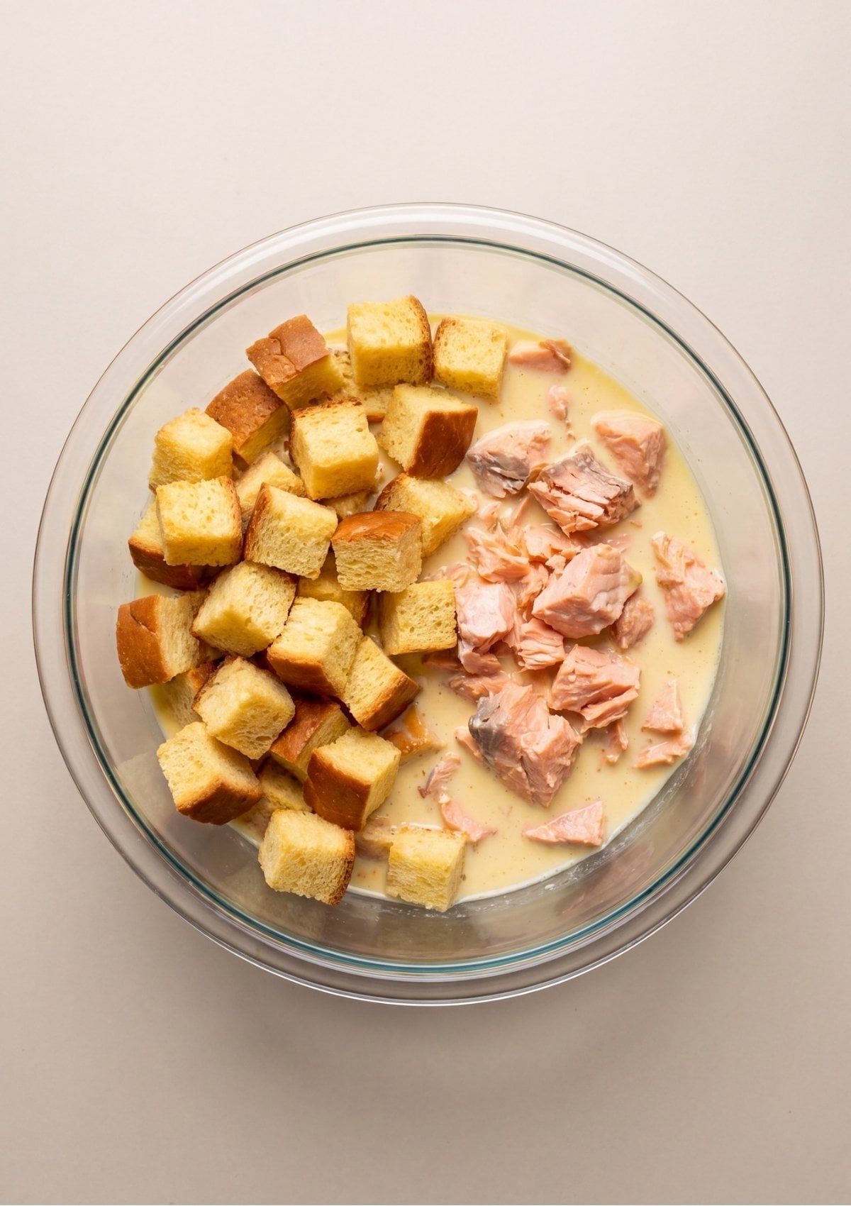 Cubed brioche bread and flaked pink salmon sitting in a clear glass mixing bowl filled with an egg and milk mixture before being folded together