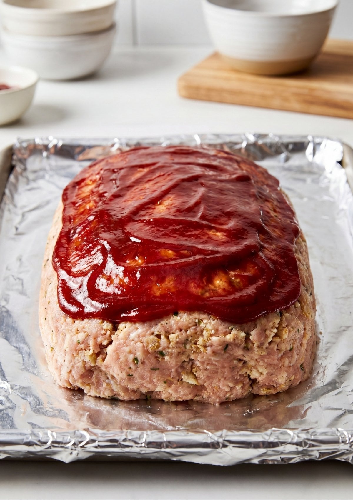 Raw turkey meatloaf on a foil-lined tray being topped with a thick layer of red tomato-based glaze.