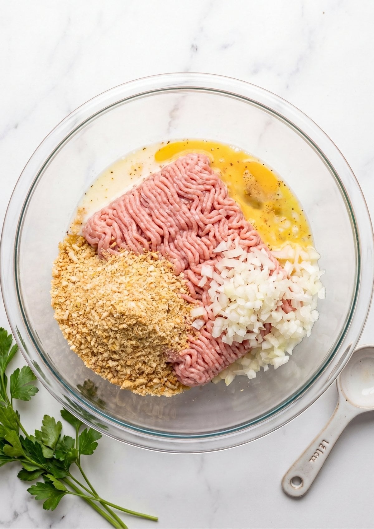 A clear glass bowl containing raw ground turkey, breadcrumbs, and finely diced white onions being added to a whisked egg mixture.