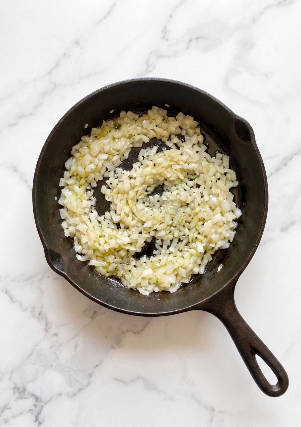 Finely chopped onions cooking with olive oil in a black cast iron skillet on a white marble countertop