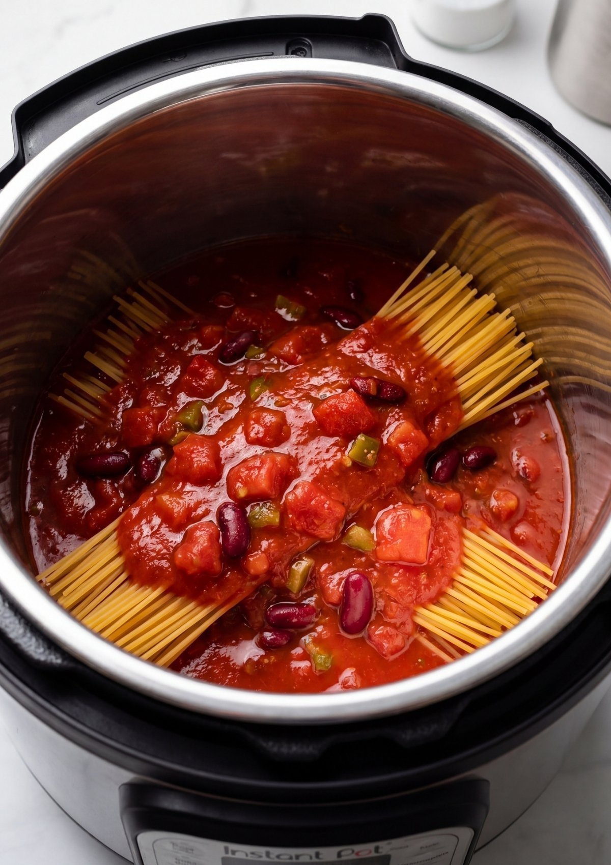 Dry spaghetti noodles arranged in a crisscross pattern inside an Instant Pot, topped with chunky tomato sauce and kidney beans.