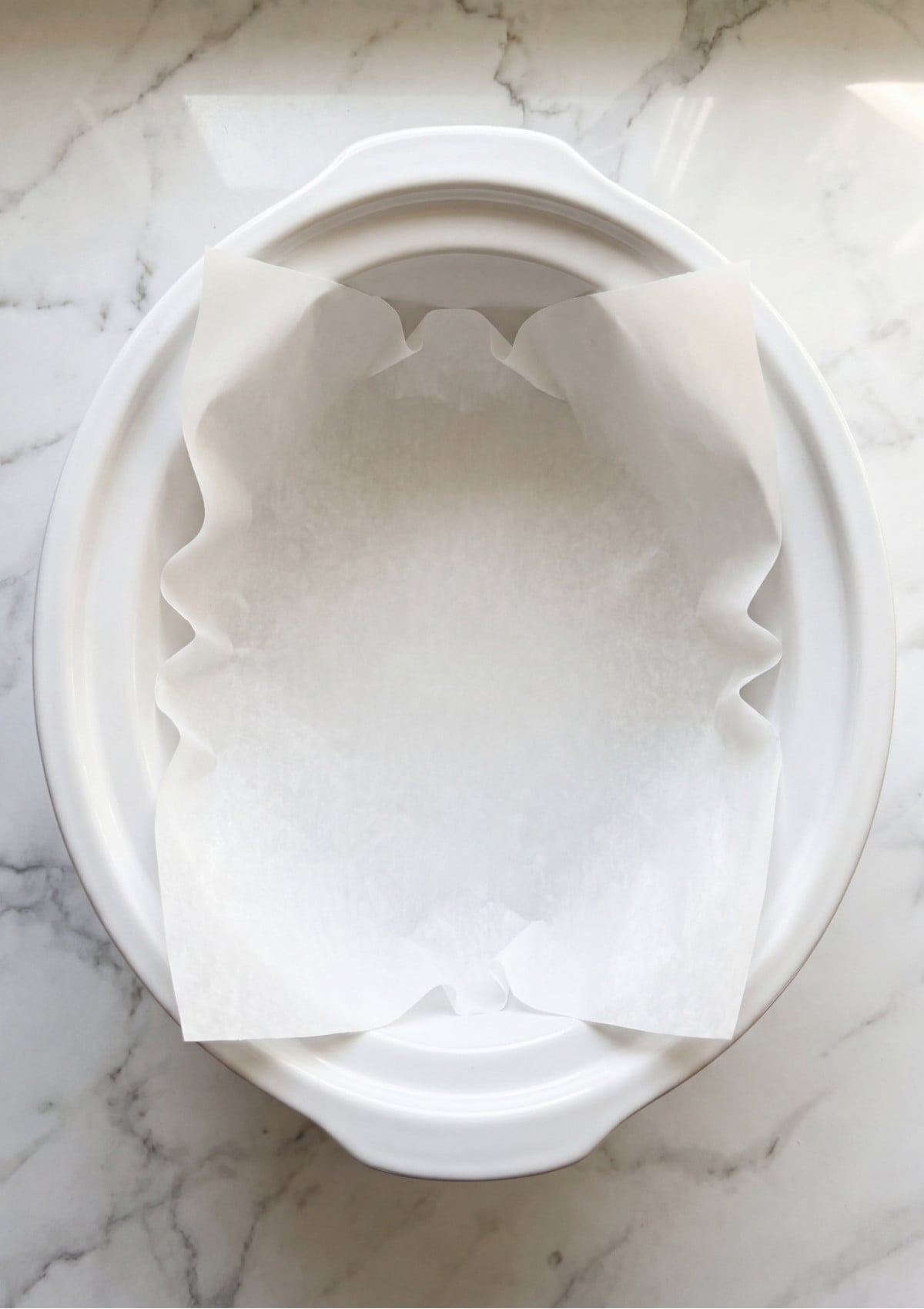 An empty white ceramic slow cooker bowl lined with a clean sheet of white parchment paper