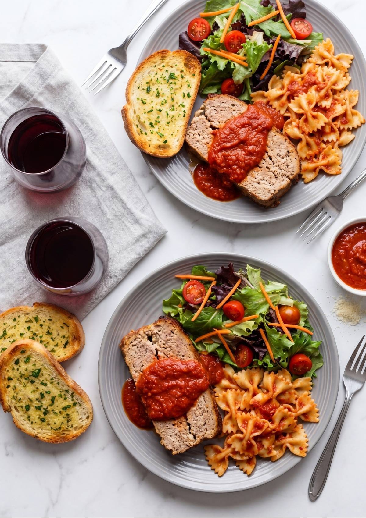 Two plates served with slices of turkey meatloaf topped with marinara sauce, bow tie pasta, side salads, and garlic bread on a white marble surface with two glasses of red wine