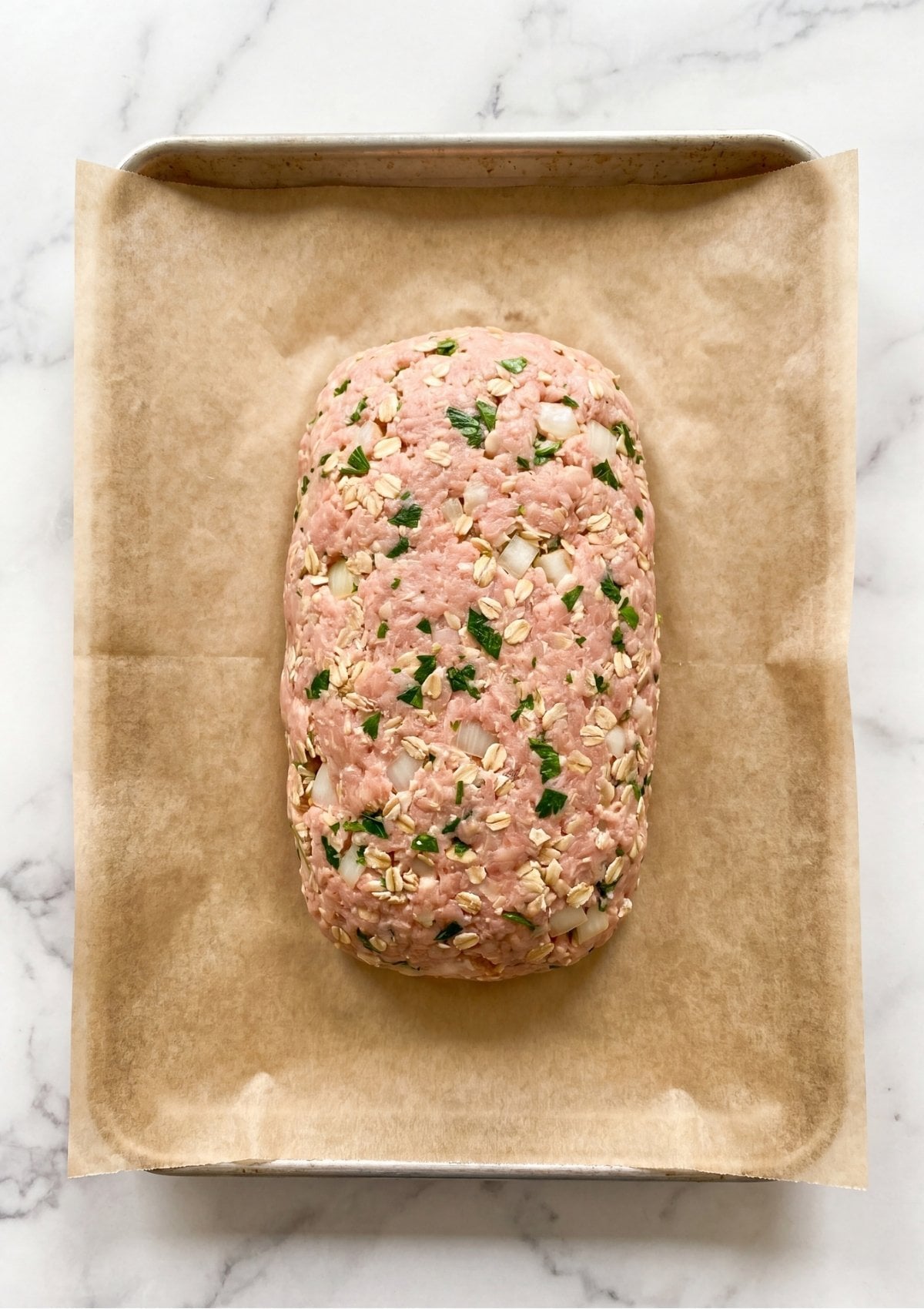 Uncooked ground turkey meatloaf mixture containing visible oats, onions, and parsley shaped into a neat loaf on parchment paper.