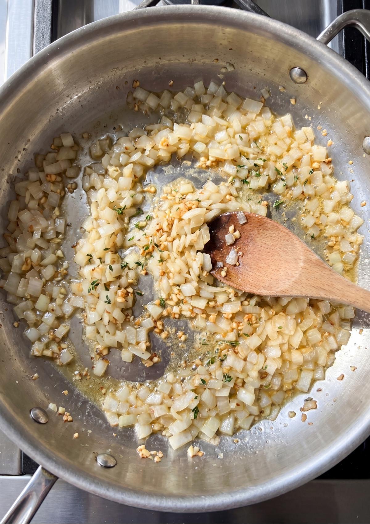 Diced yellow onions, minced garlic, and fresh thyme cooking in a stainless steel skillet with a wooden spoon stirring the mixture