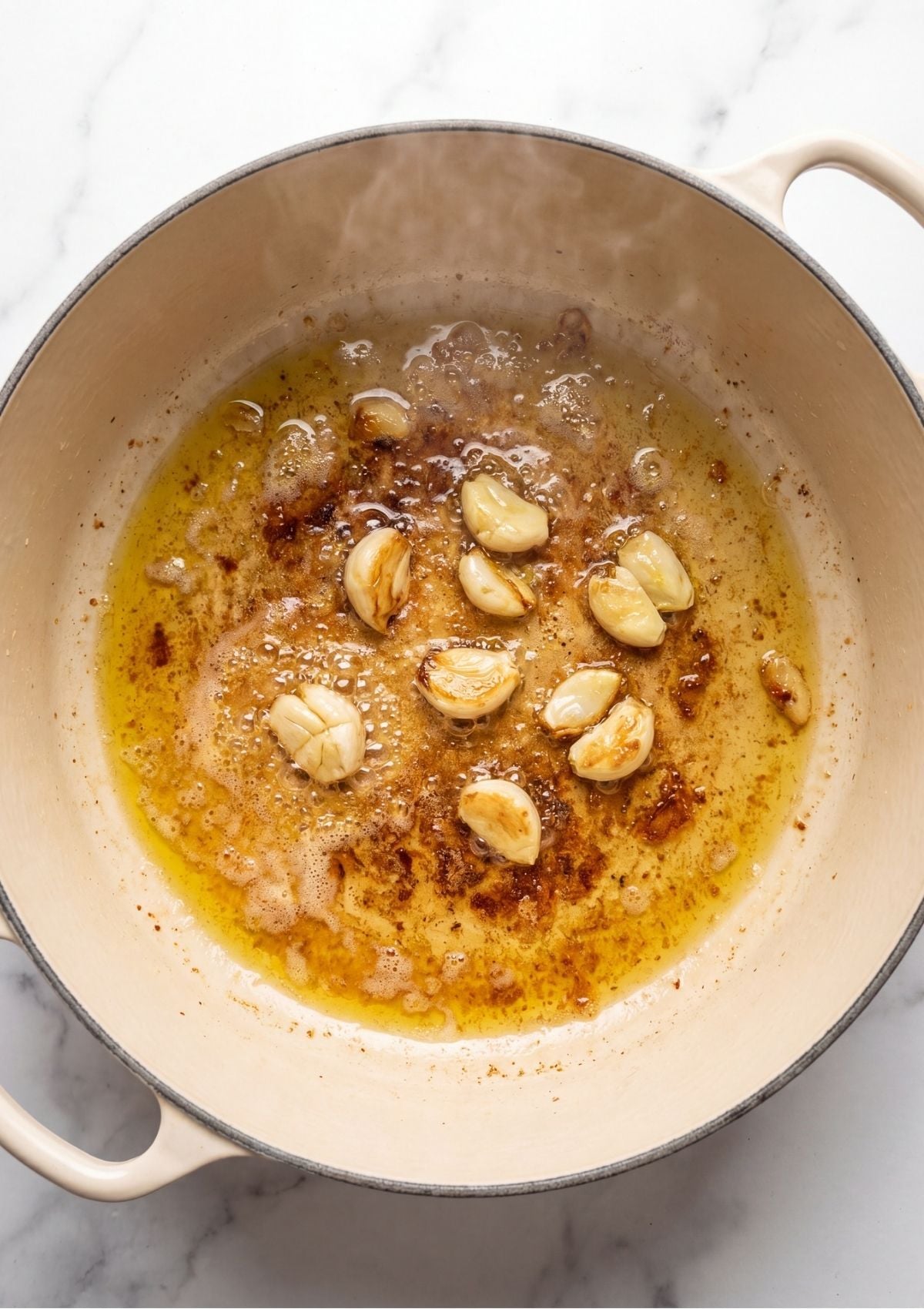 Whole garlic cloves cooking and bubbling in pan drippings and oil inside a hot white Dutch oven.