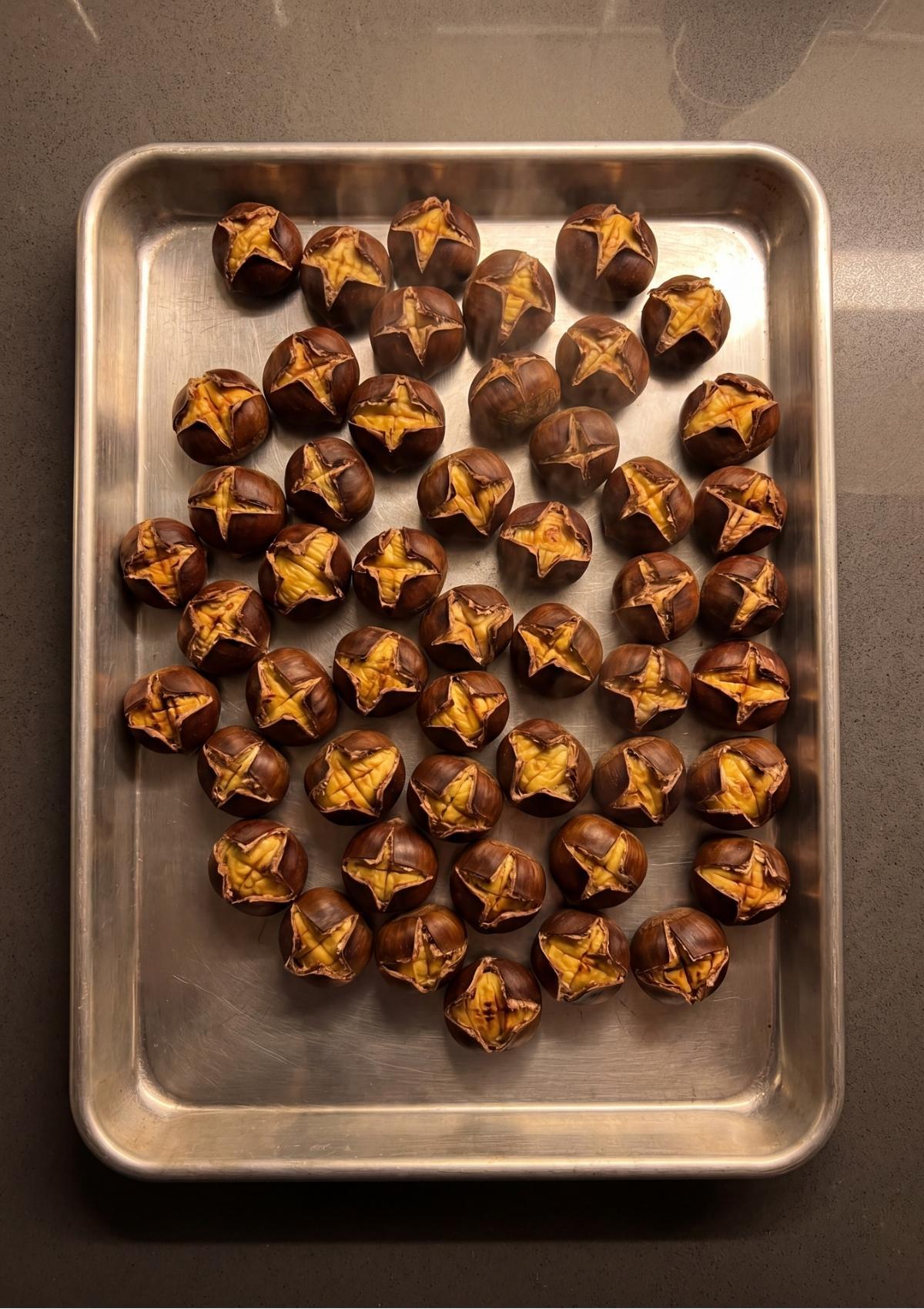 A metal baking sheet filled with roasted chestnuts in their shells, all curled open from the oven heat.