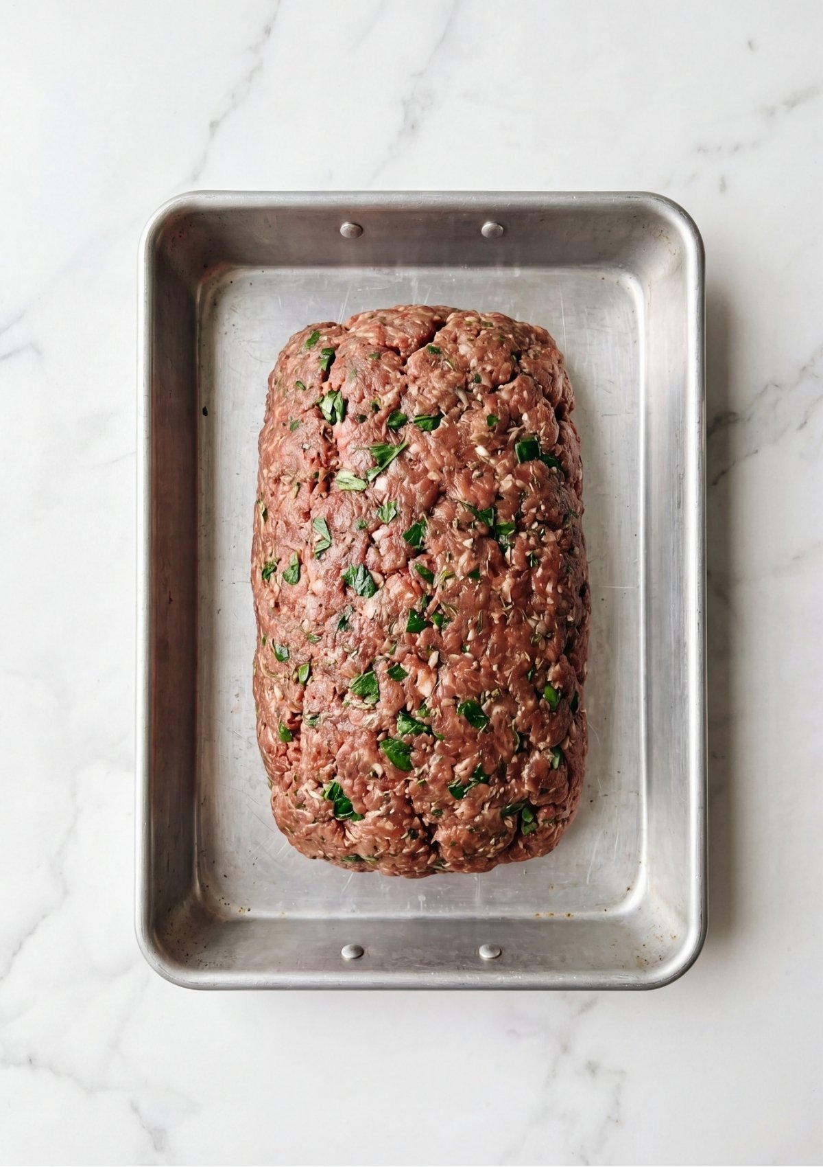Raw ground meat mixture containing visible green herbs and onions shaped into a loaf in a silver baking pan.