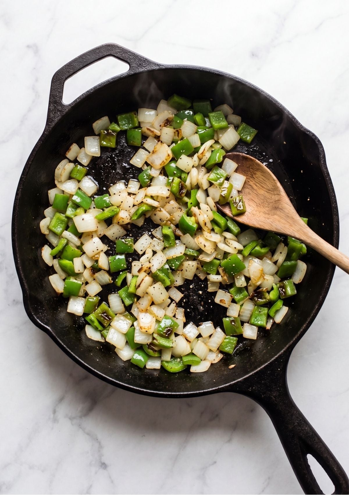 Diced green bell peppers and white onions being softened in a black cast-iron skillet with a wooden spoon.
